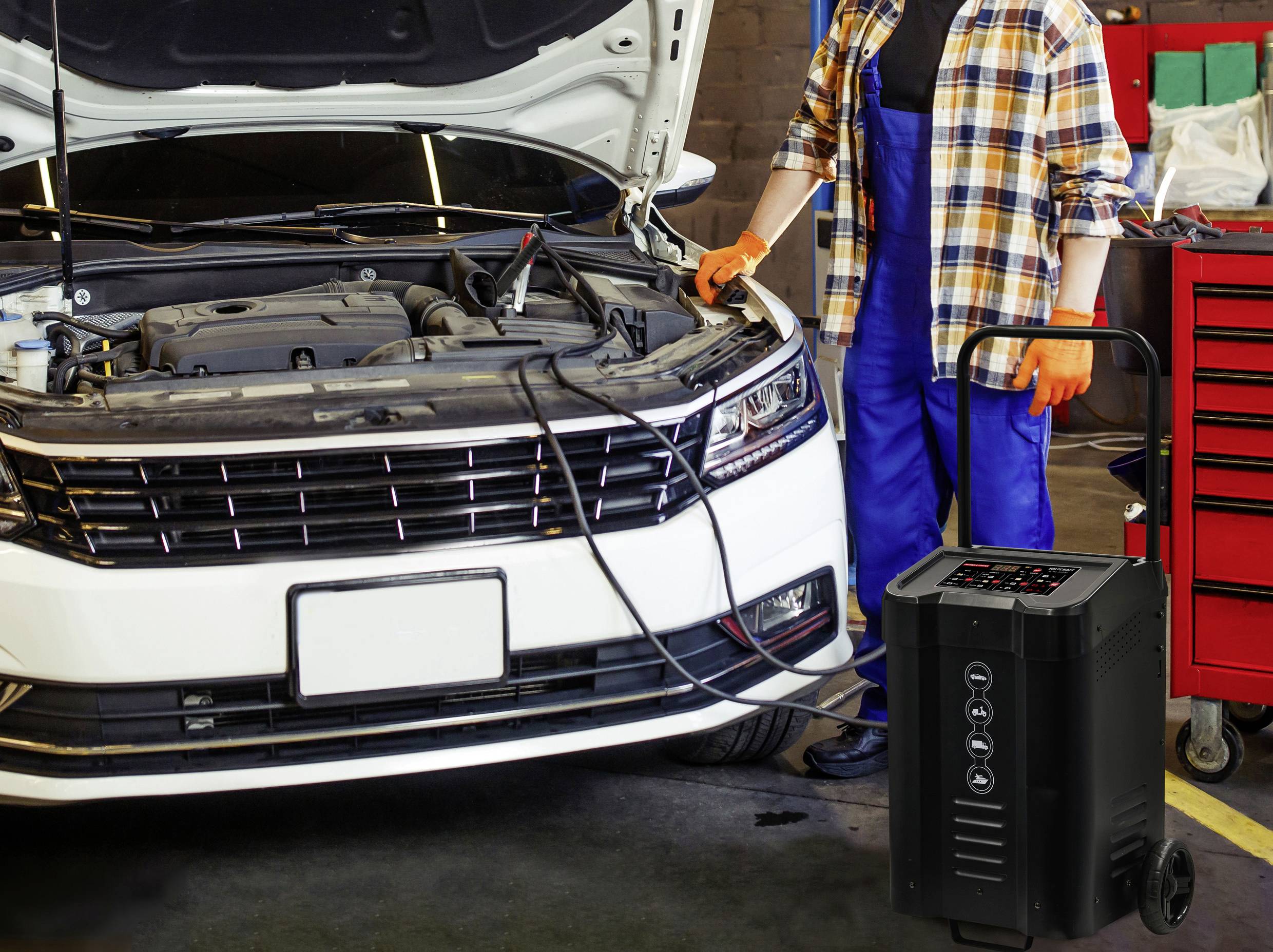 Un mécanicien se tient à côté d'une voiture dont le capot est ouvert. À ses côtés se trouve un chargeur de batterie, relié à la batterie du véhicule.