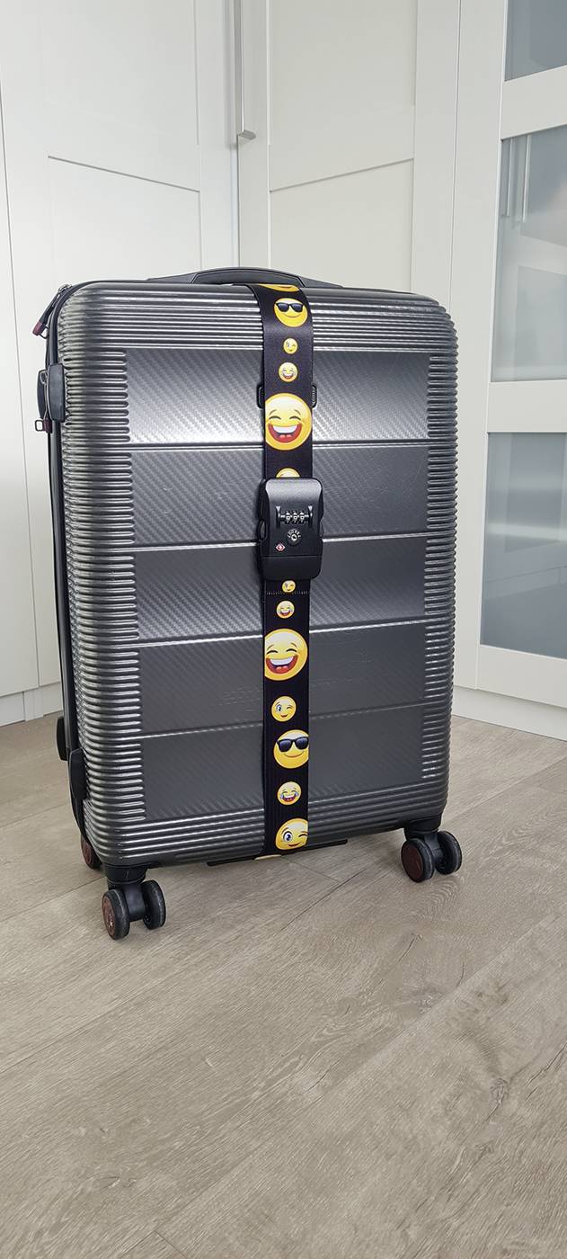Une valise noire à quatre roues avec une sangle de bagage à motif emoji, posée sur un plancher en bois devant un placard blanc.