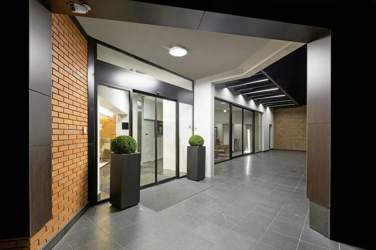 Entrée d'un bâtiment moderne avec une façade vitrée, des briques rouges et deux plantes dans des conteneurs carrés à l'entrée.