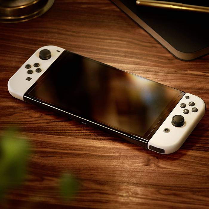 Une console de jeu portable est posée sur une table en bois. Elle possède des manettes amovibles de chaque côté. Un ordinateur portable est visible en arrière-plan.