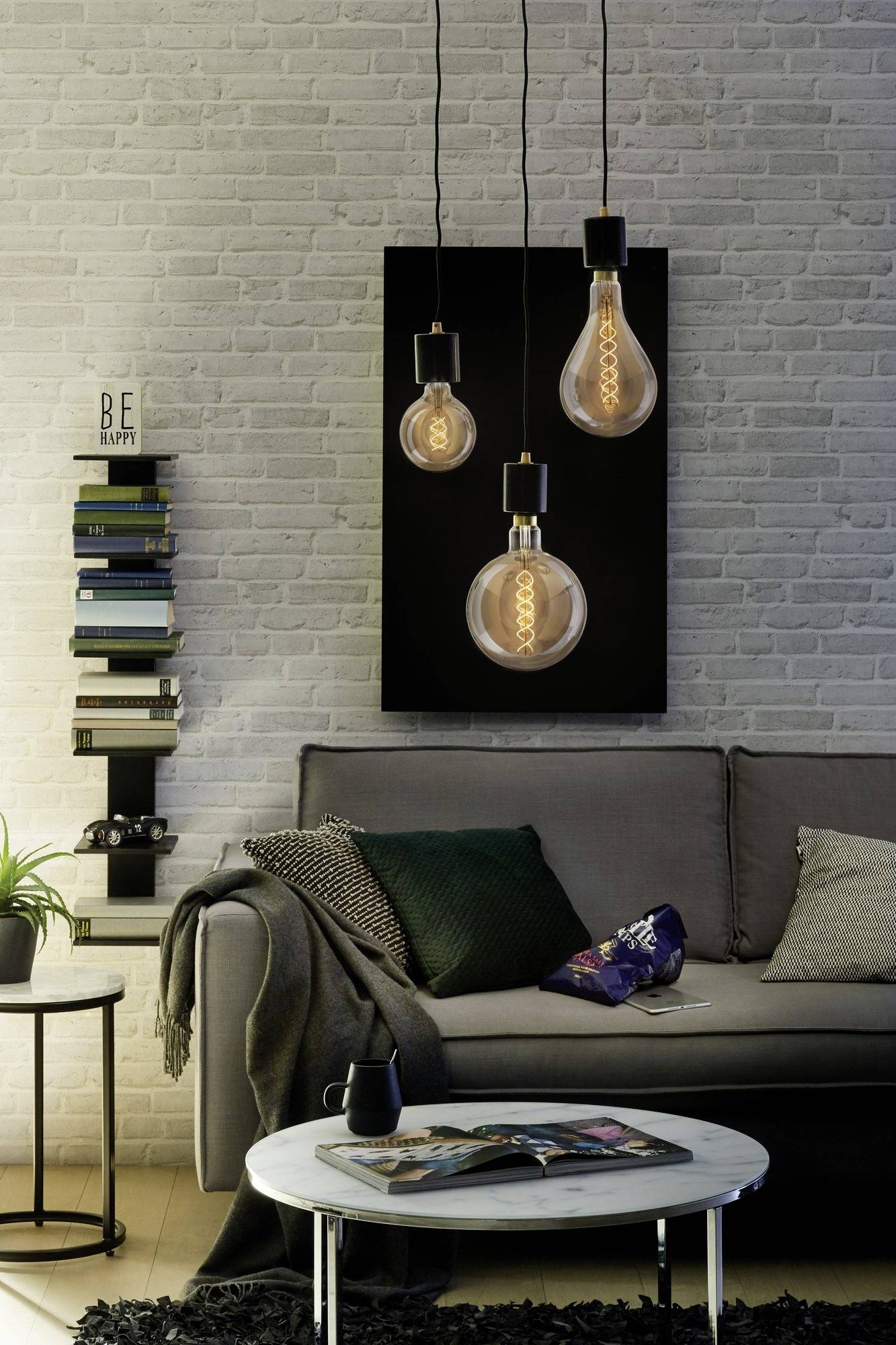 Décoration de salon moderne : Canapé gris avec des coussins verts et bleus, trois lampes suspendues en forme d'ampoules, pile de livres contre le mur.