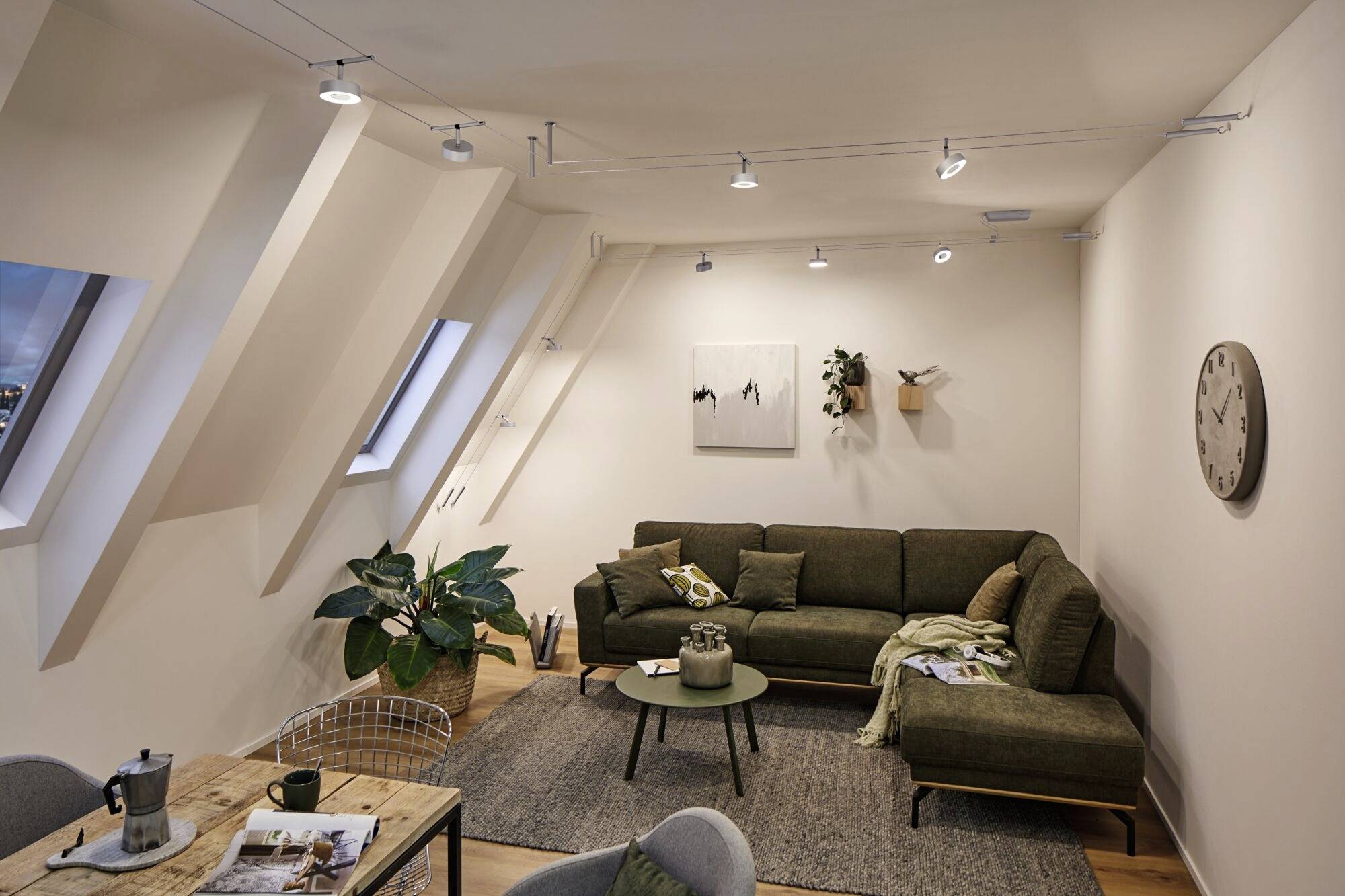 Un salon confortable avec un canapé d'angle vert, des tableaux accrochés aux murs et une table basse au centre. Une horloge est suspendue à droite.