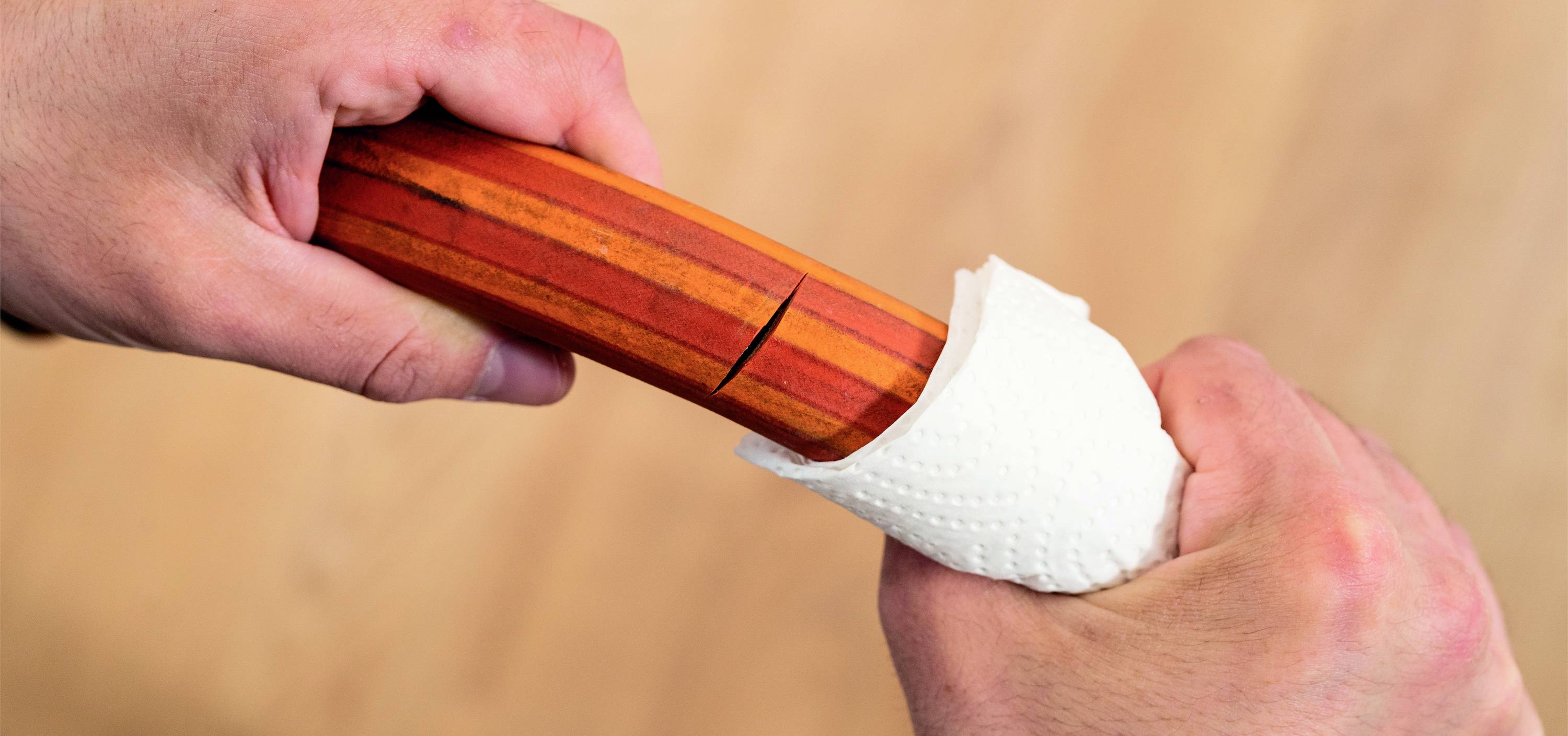 Deux mains saisissent un bâton en bois rouge portant un pochoir en papier blanc, sur un fond brun clair.