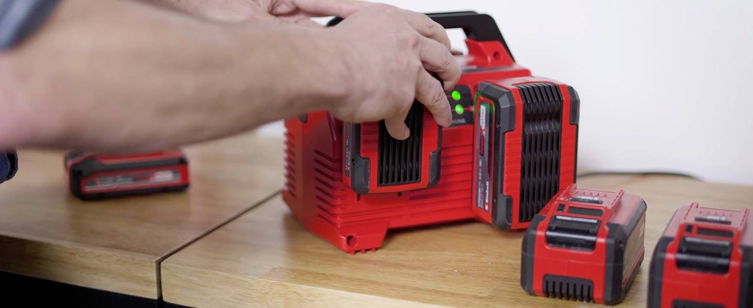 Une personne insère des batteries rechargeables rouges dans un chargeur posé sur une table en bois. Des voyants verts indiquent l'état de charge.