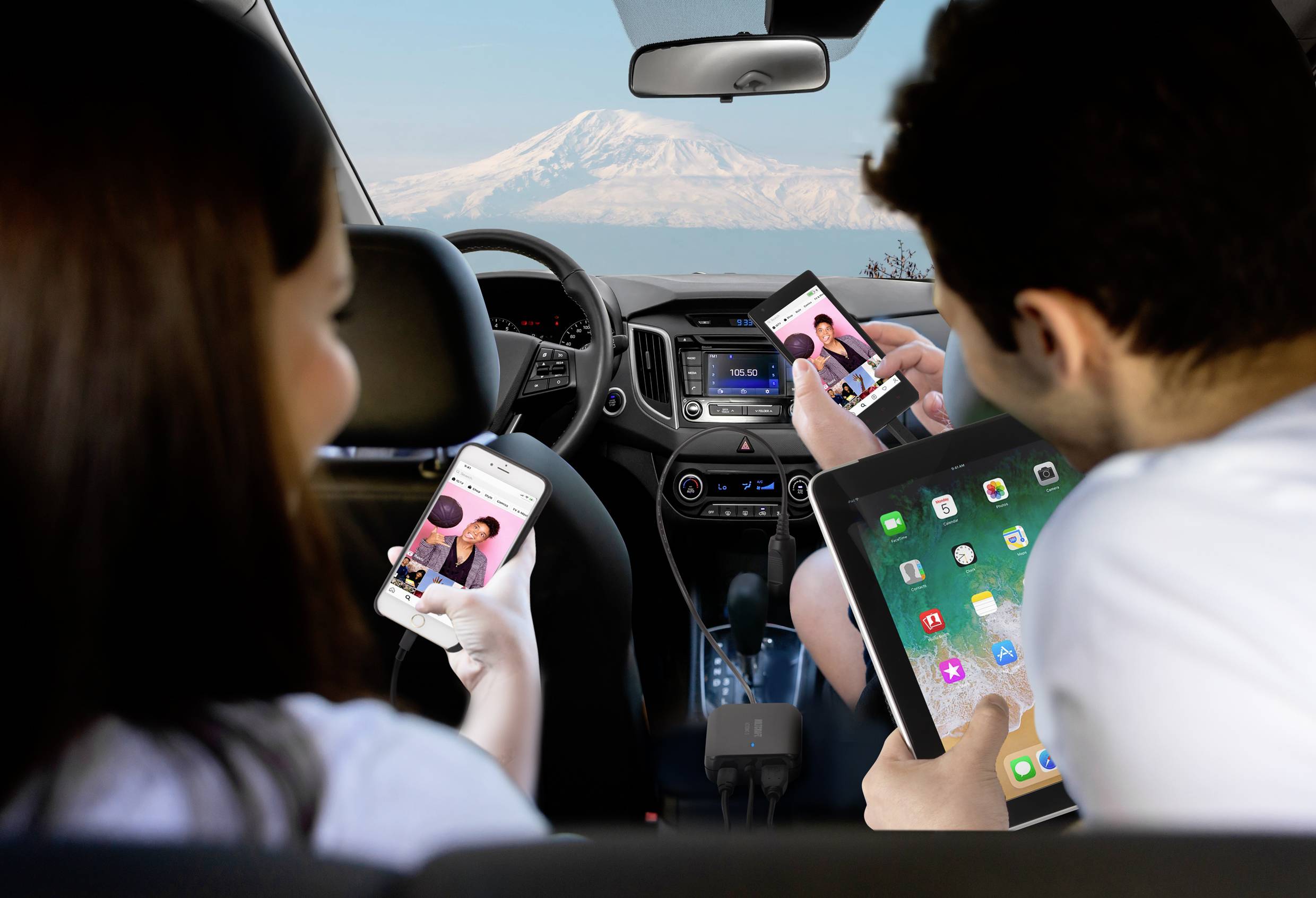 Un homme et une femme sont assis dans une voiture et utilisent leurs smartphones, tandis qu'une tablette est posée sur les genoux de l'homme. À l'arrière-plan, on aperçoit une montagne enneigée.