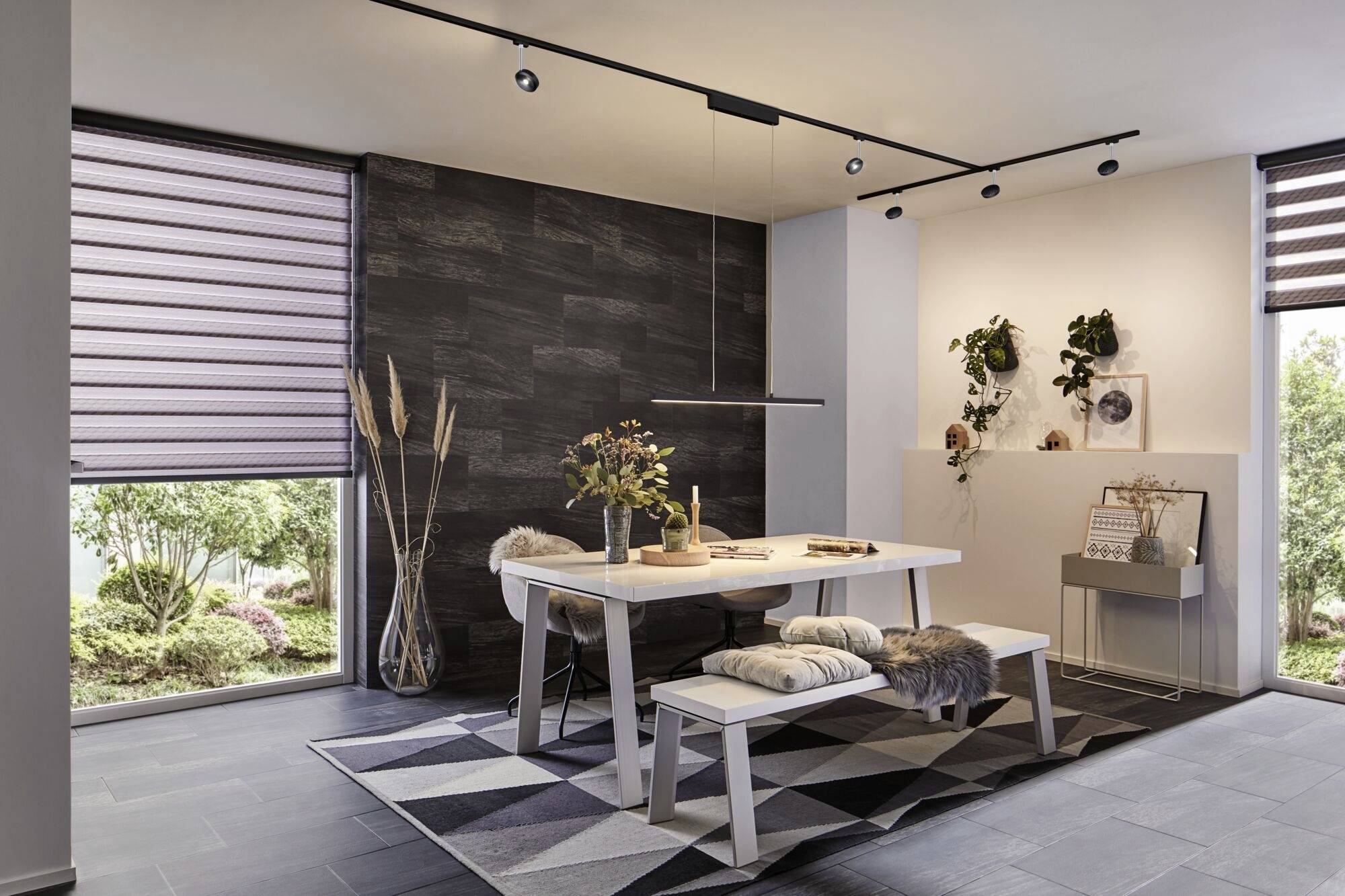 Salle à manger avec une table moderne et un banc sur un tapis géométrique. Mur sombre, plantes décoratives et grandes fenêtres avec stores.
