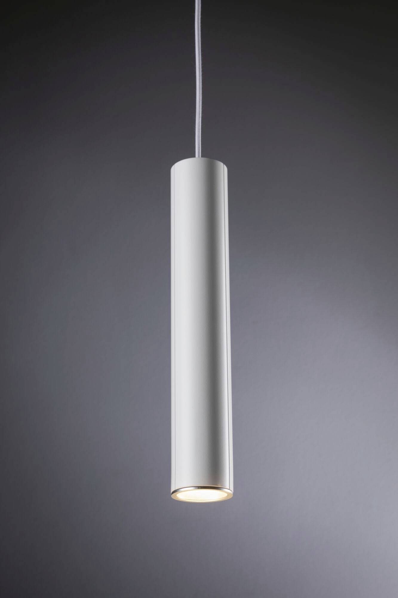 Une lampe suspendue élancée, cylindrique en métal, suspendue dans un espace moderne aux tons neutres. Elle éclaire vers le bas.