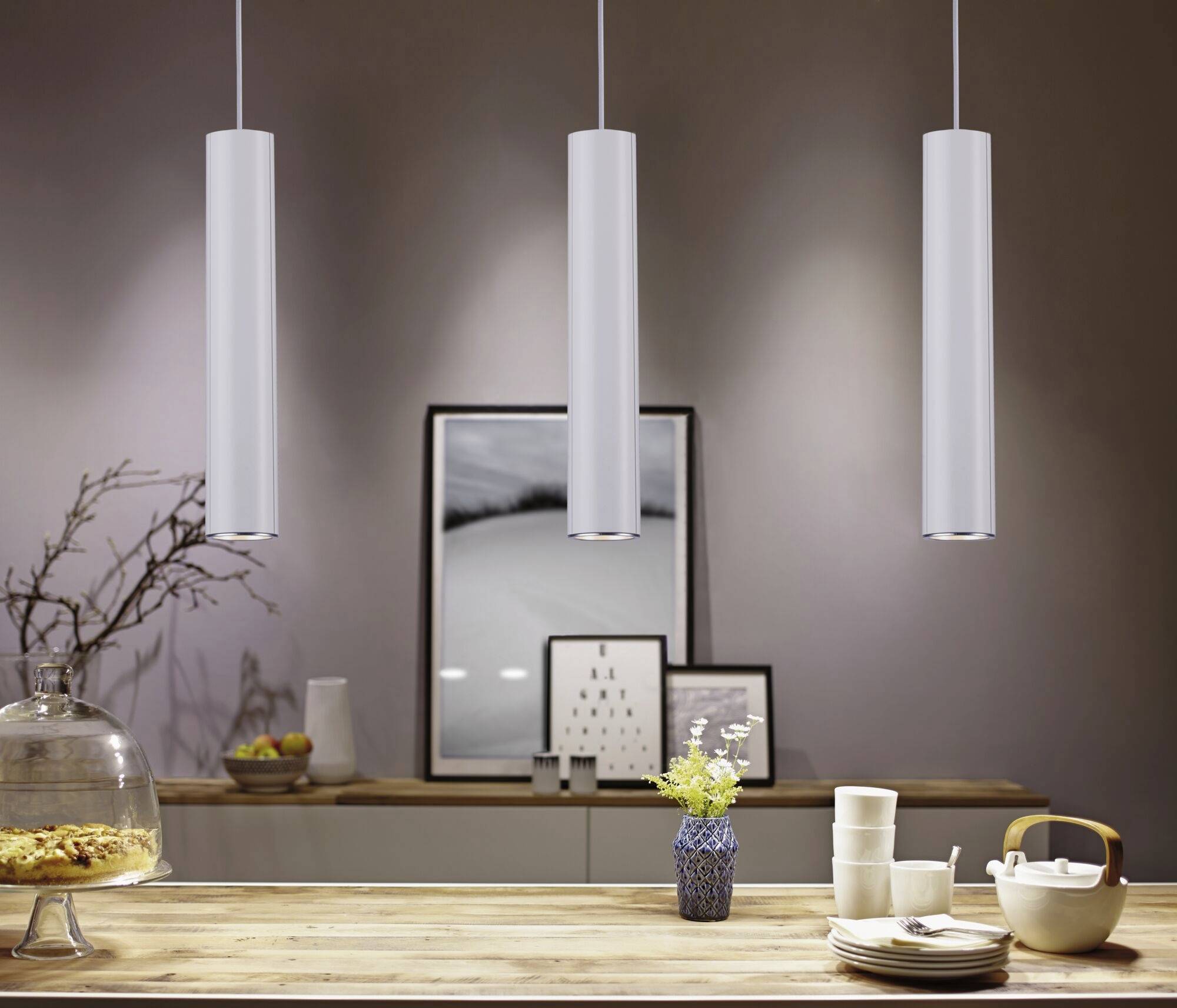 Espace repas moderne avec trois lampes suspendues au-dessus d'une table décorée avec de la vaisselle et un vase. Une œuvre d'art est accrochée au mur en arrière-plan.