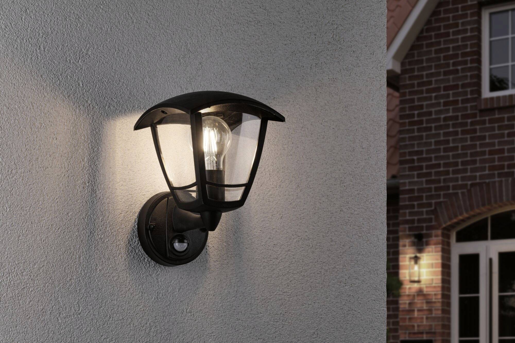 Lampe extérieure fixée sur un mur de maison, allumée au crépuscule. À l'arrière-plan, une maison de briques avec une fenêtre éclairée.