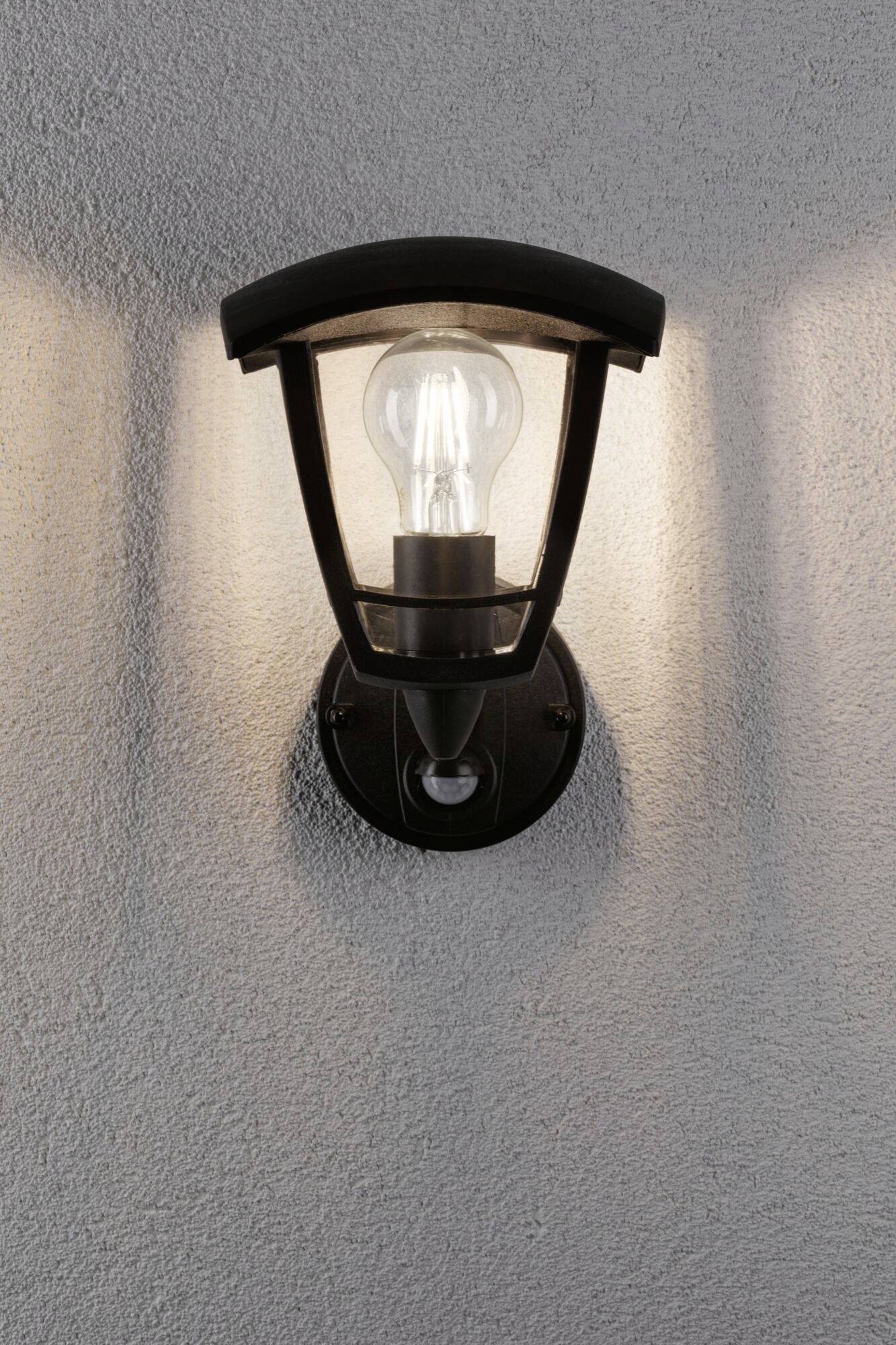 Applique murale extérieure avec un boîtier noir moderne, fixée sur un mur gris texturé. La lampe est allumée et diffuse une lumière chaleureuse.