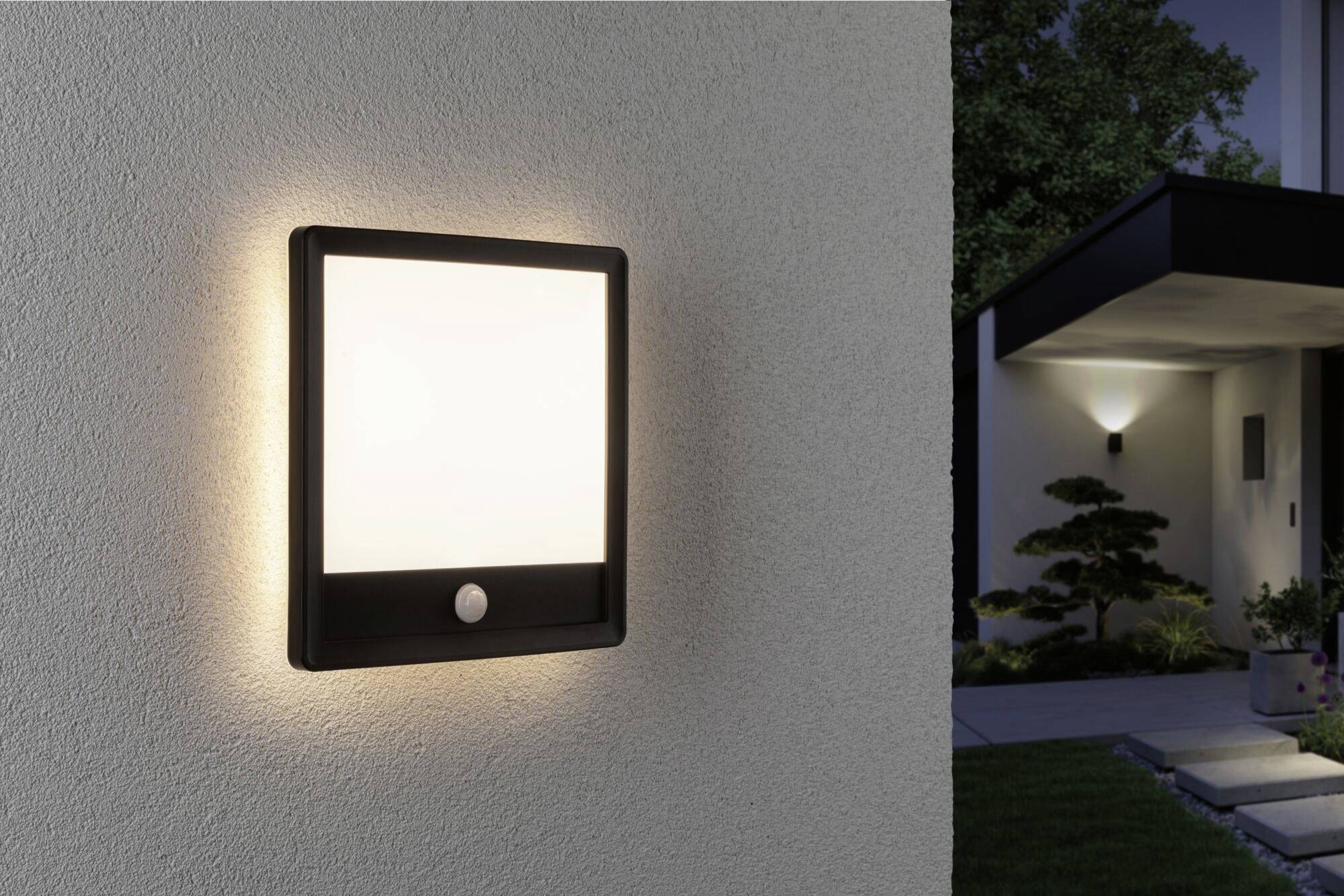 Lampe extérieure avec détecteur de mouvement sur le mur de la maison, illuminant la zone d'entrée. À l'arrière-plan, on peut voir une porte d'entrée moderne.