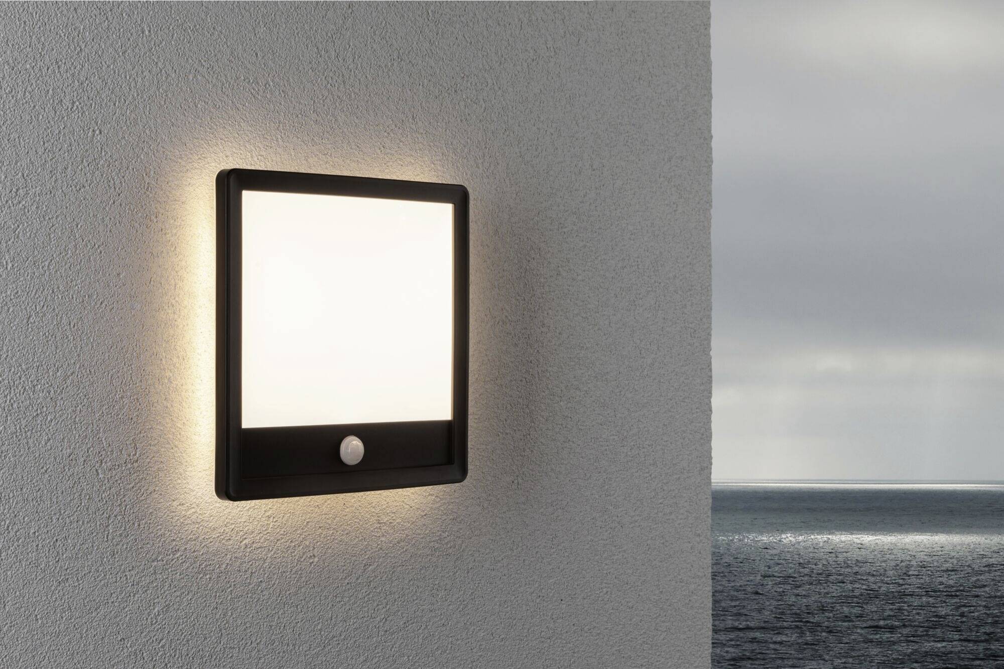 Une lampe extérieure carrée est montée sur le mur. Elle s'allume à la tombée du jour, la mer est visible en arrière-plan.
