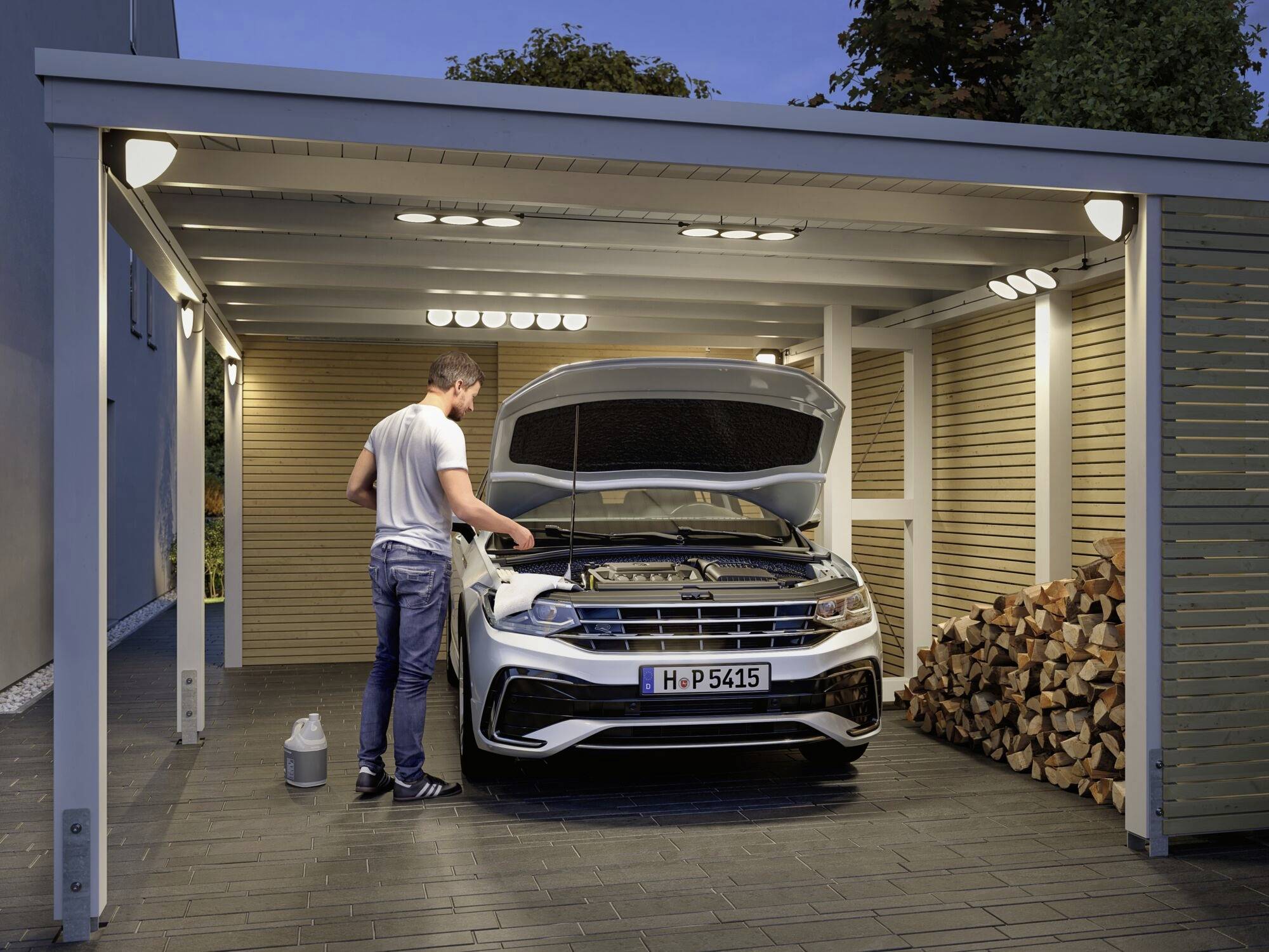 Un homme vérifie le moteur d'une voiture blanche sous un carport ouvert. À côté, un tas de bois de chauffage est empilé.