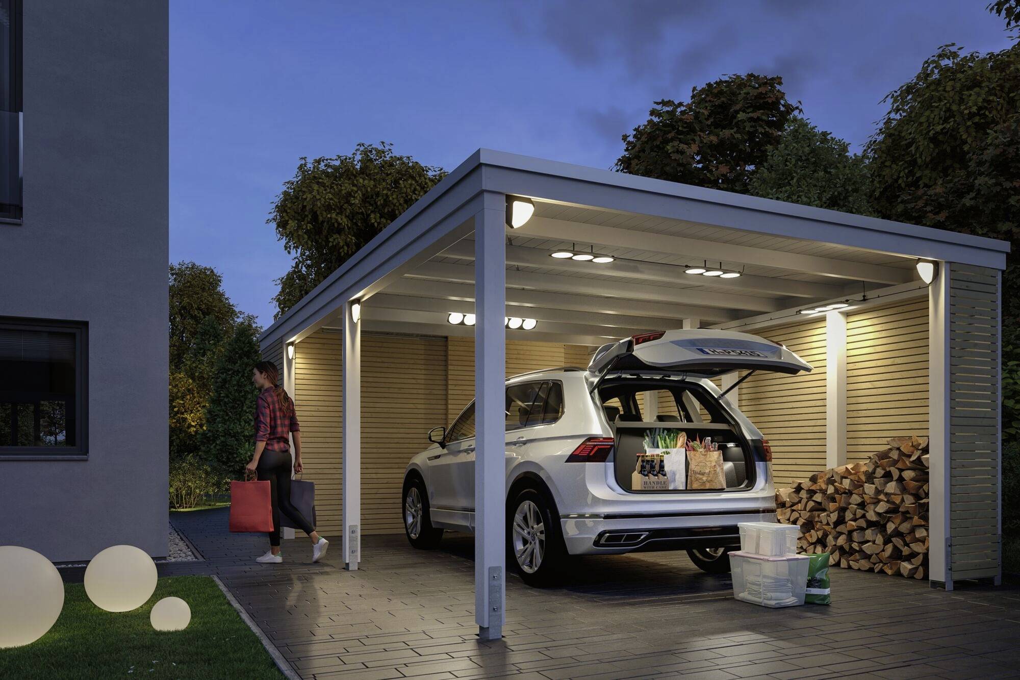 Une personne transporte des bagages vers un carport éclairé et couvert, avec un coffre ouvert rempli de courses.
