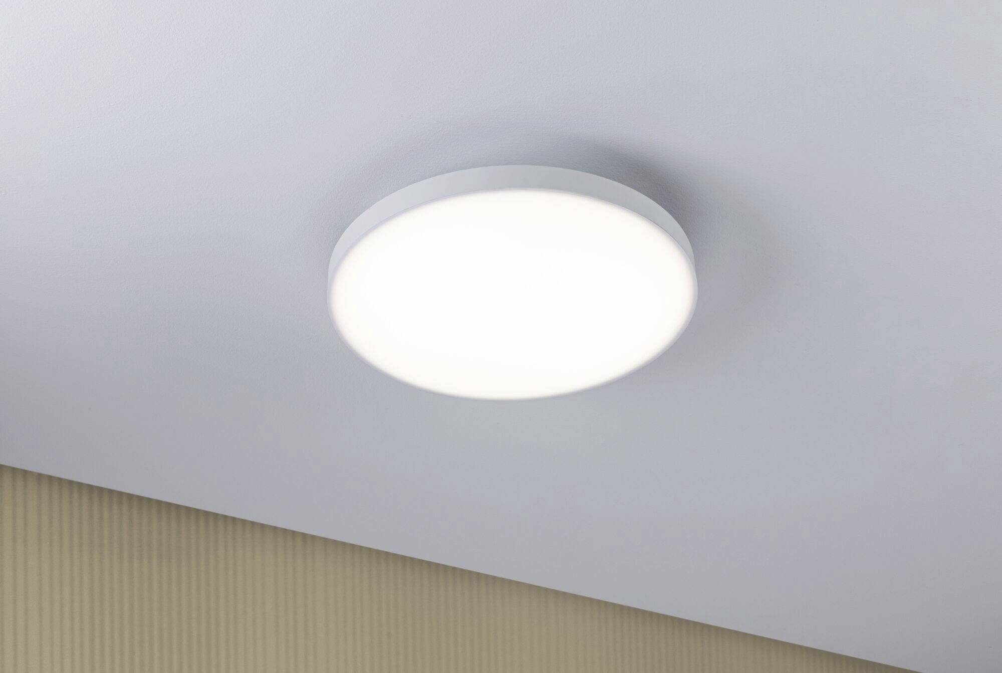 Plafonnier rond blanc, fixé à un plafond gris, qui diffuse une lumière uniforme. En dessous, une partie d'un mur peint en beige est visible.