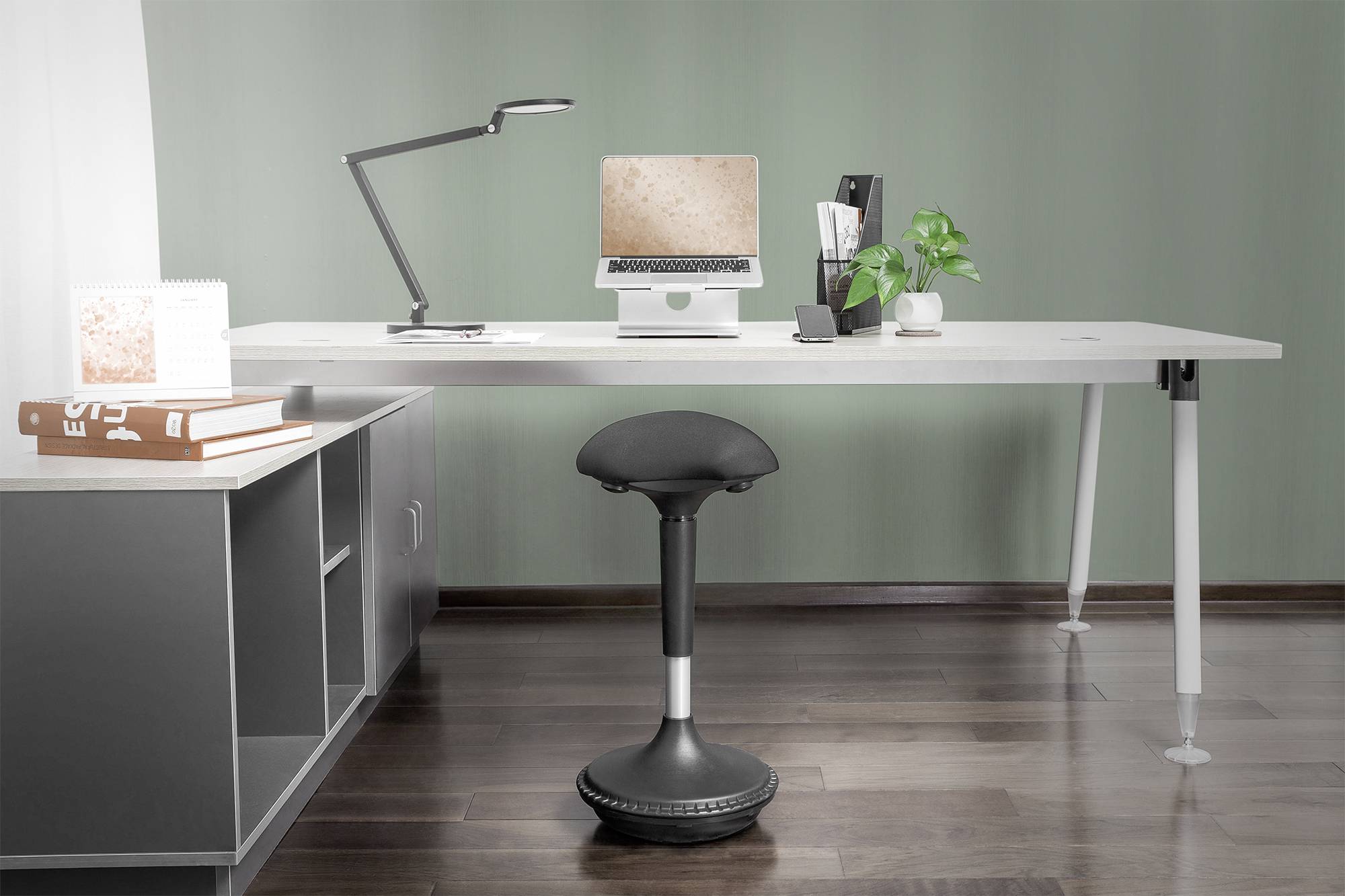 Bureau moderne avec un ordinateur portable sur un support, une lampe de bureau, une plante et un carnet de notes. Au premier plan se trouve un tabouret ergonomique.