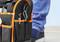Un sac à outils noir avec des coutures orange est posé sur un sol. Une personne portant un jean bleu et des baskets se tient à côté.