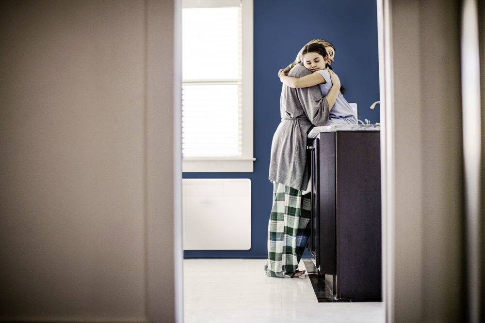 Une personne en pyjama enlace une autre dans la cuisine. Ils sont debout près de l'évier. En arrière-plan, un mur bleu et une fenêtre sont visibles.