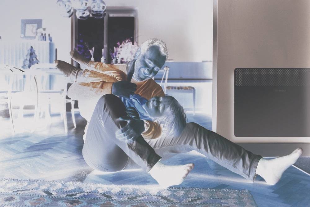 Un homme est assis par terre et enlace ludiquement un enfant. Ils rient ensemble, dans un salon avec un tapis et des meubles.