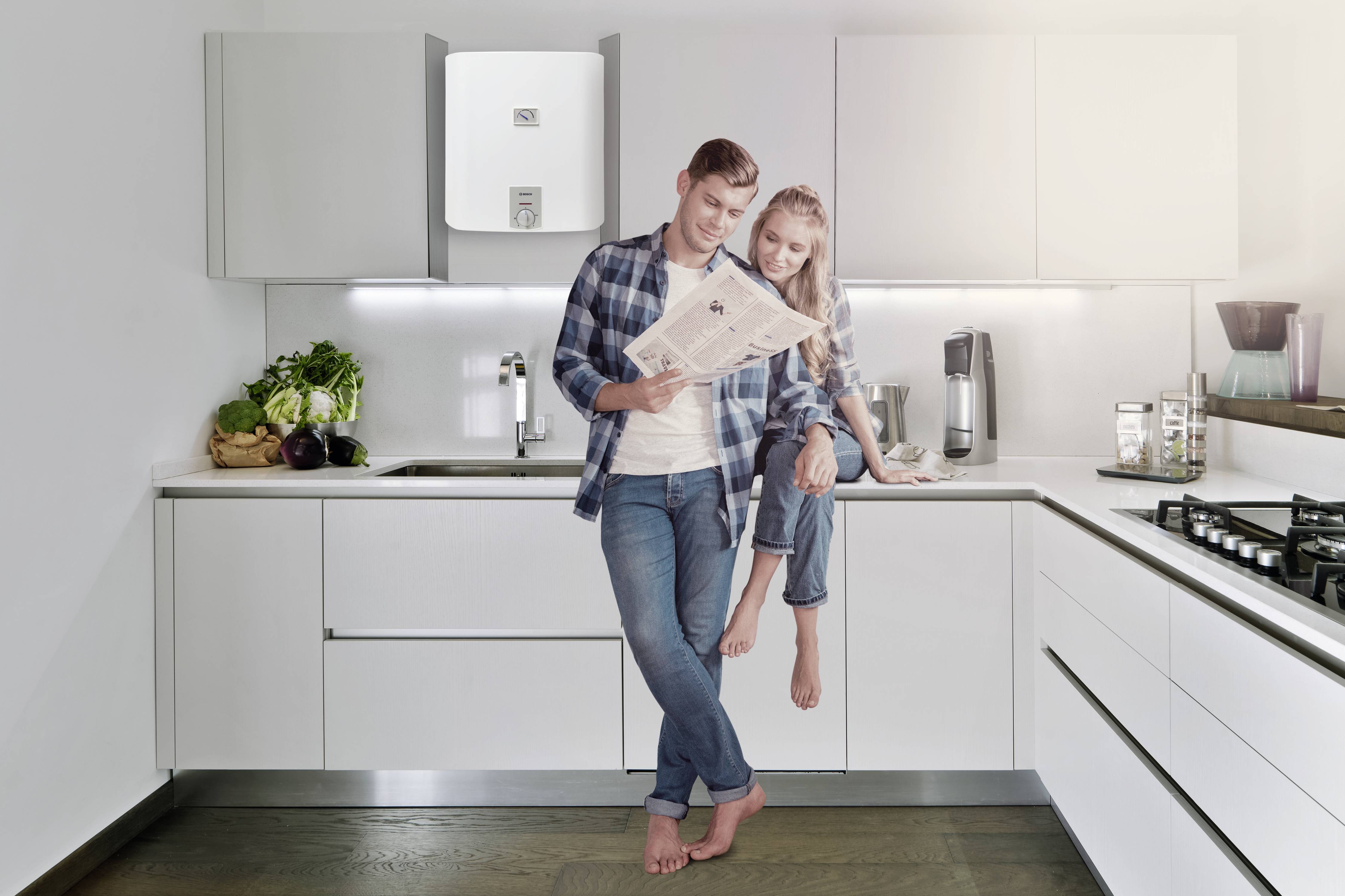 Un couple souriant se tient dans une cuisine moderne. L'homme lit un journal, tandis que la femme est assise sur le plan de travail. Des légumes frais sont visibles en arrière-plan.