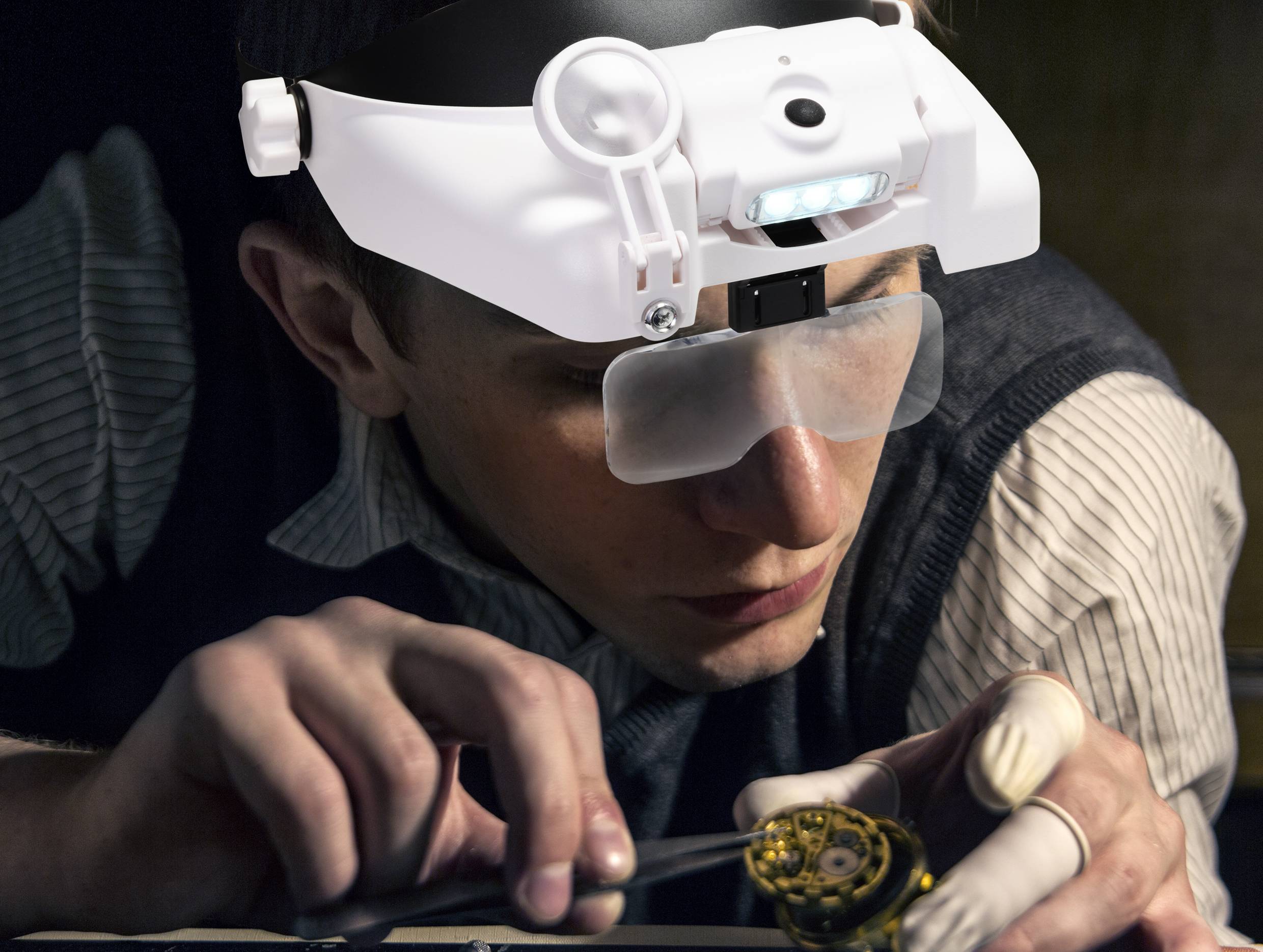 Une personne porte des lunettes loupe avec éclairage LED et travaille de manière concentrée sur un petit mécanisme d'horloge, un outil à la main.
