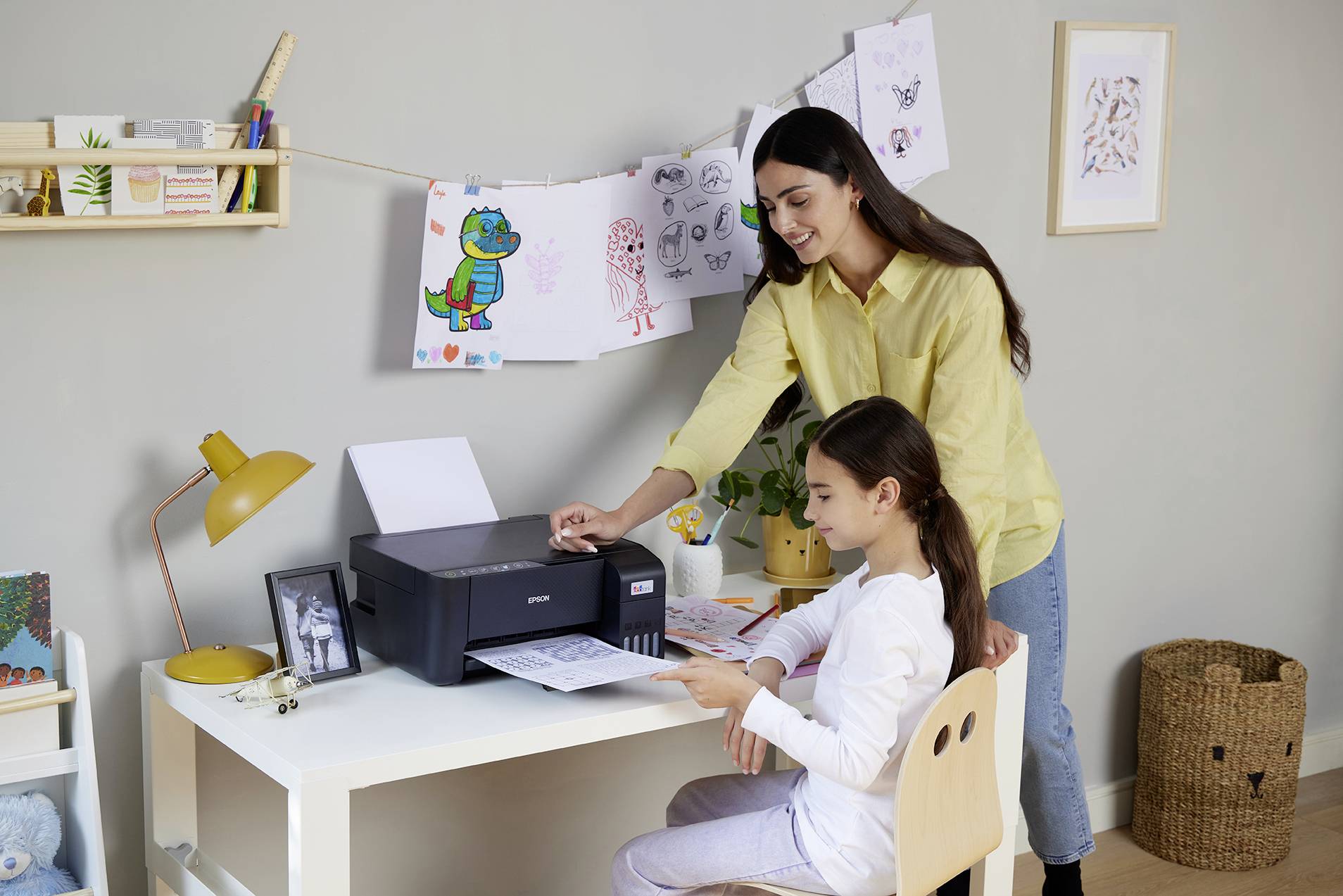 Une femme aide un enfant à imprimer quelque chose avec une imprimante. Des dessins sont suspendus à une corde à l'arrière-plan d'un salon.