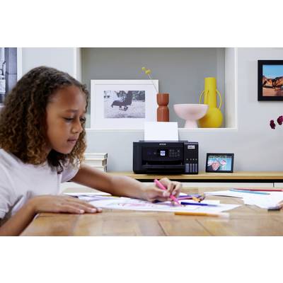 Une fille dessine avec des crayons à une table, un imprimante est posée sur une étagère en arrière-plan, avec des objets de décoration et des photos.