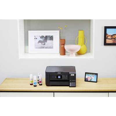 Une imprimante est posée sur une table, entourée de bouteilles d'encre. En arrière-plan, des objets décoratifs et des photos encadrées sont visibles.