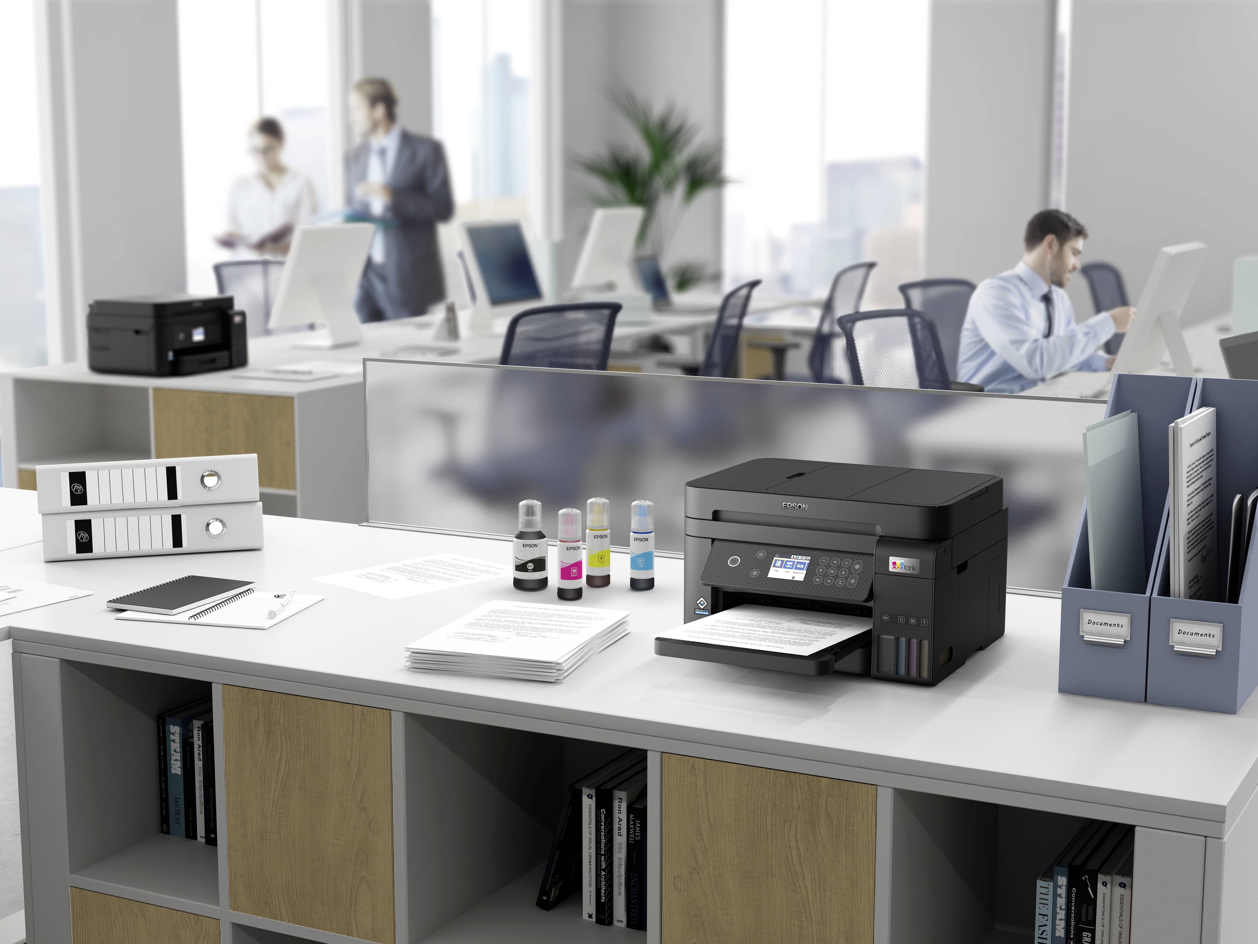 Plusieurs personnes travaillent dans un bureau moderne équipé d'ordinateurs ; au premier plan, on trouve une imprimante multifonction posée sur un bureau.