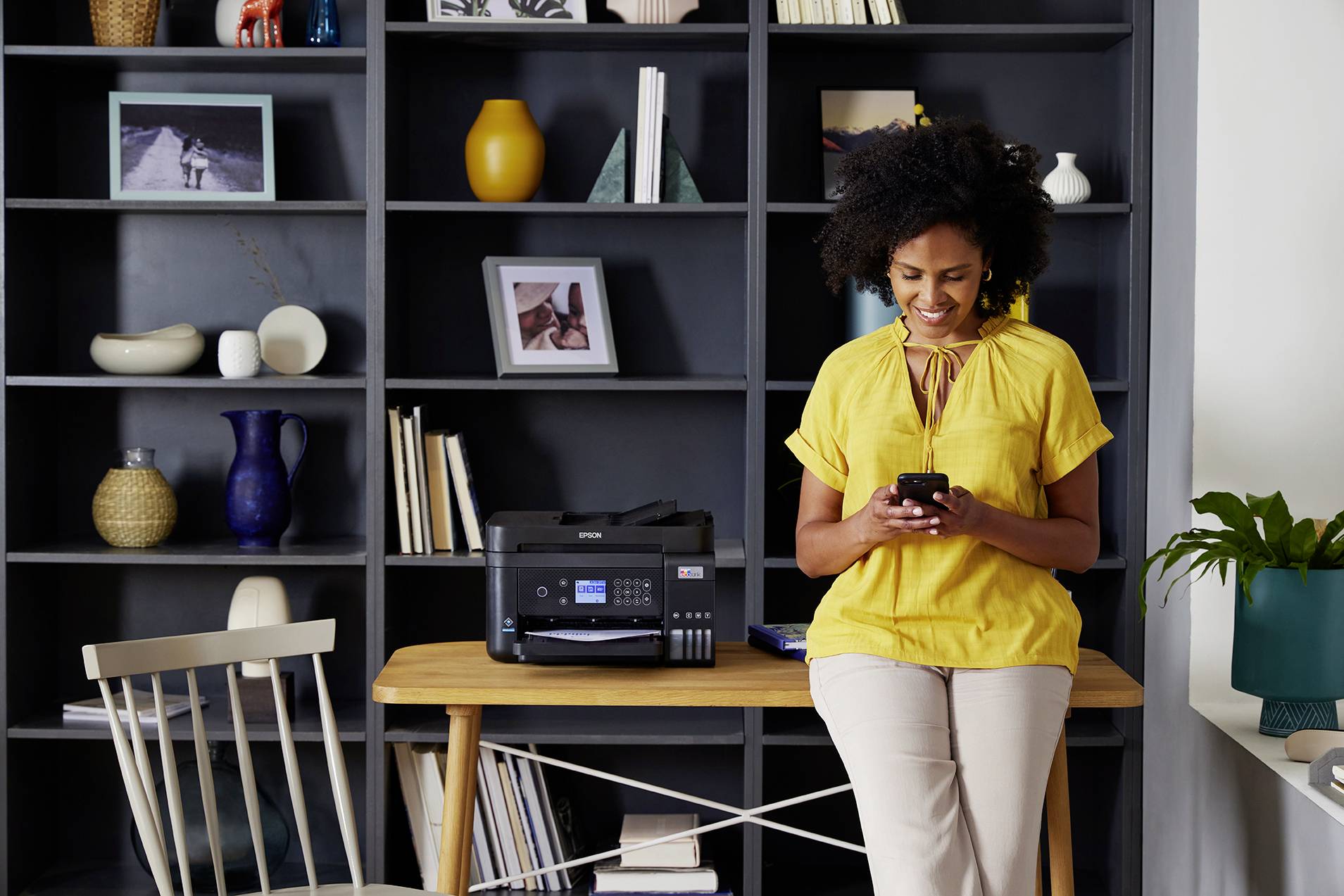 Une femme portant une blouse jaune est assise souriante avec son téléphone portable dans un bureau moderne, devant une étagère décorée et un imprimante posée sur la table.
