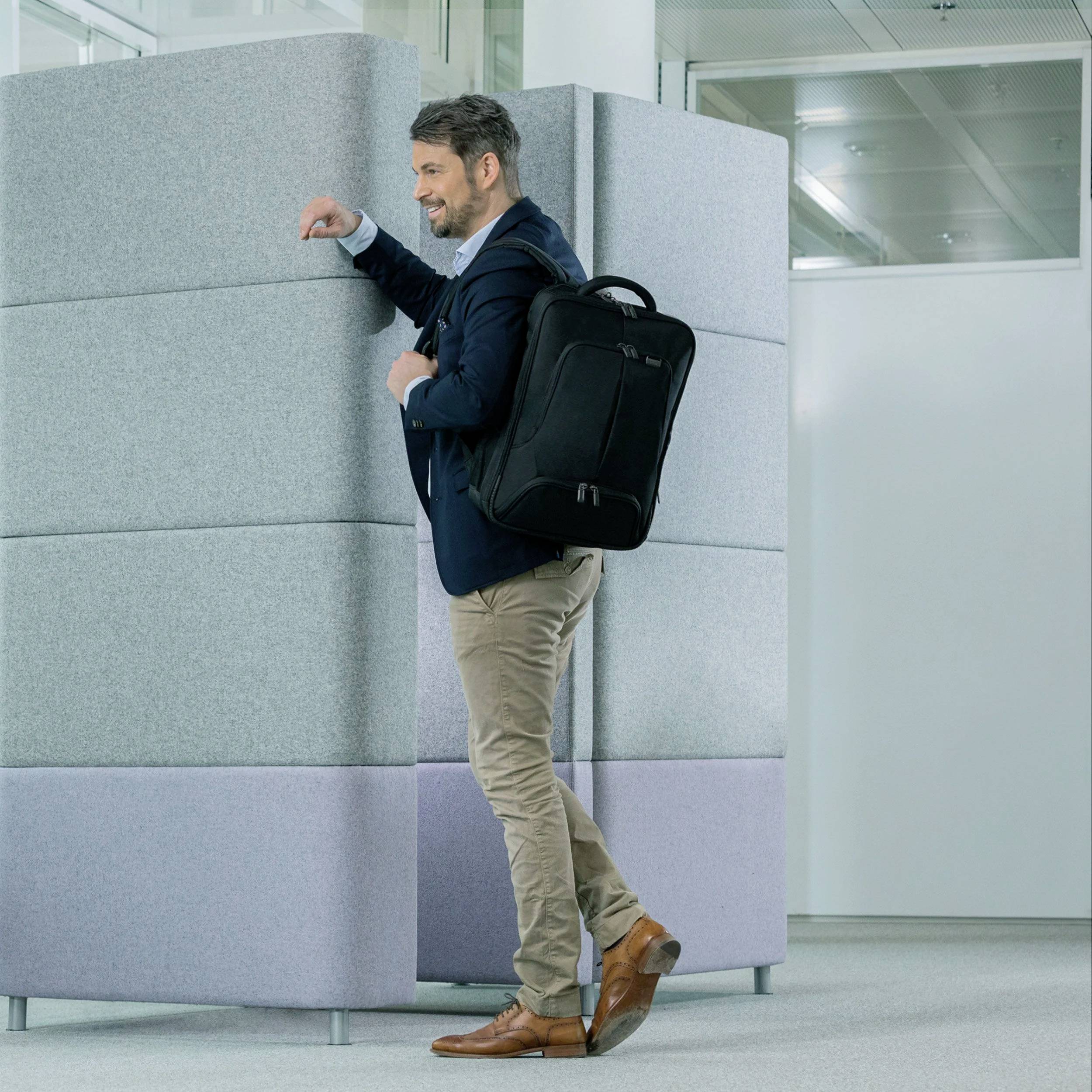 Un homme souriant portant un sac à dos traverse un espace de bureau avec une cloison grise en arrière-plan.