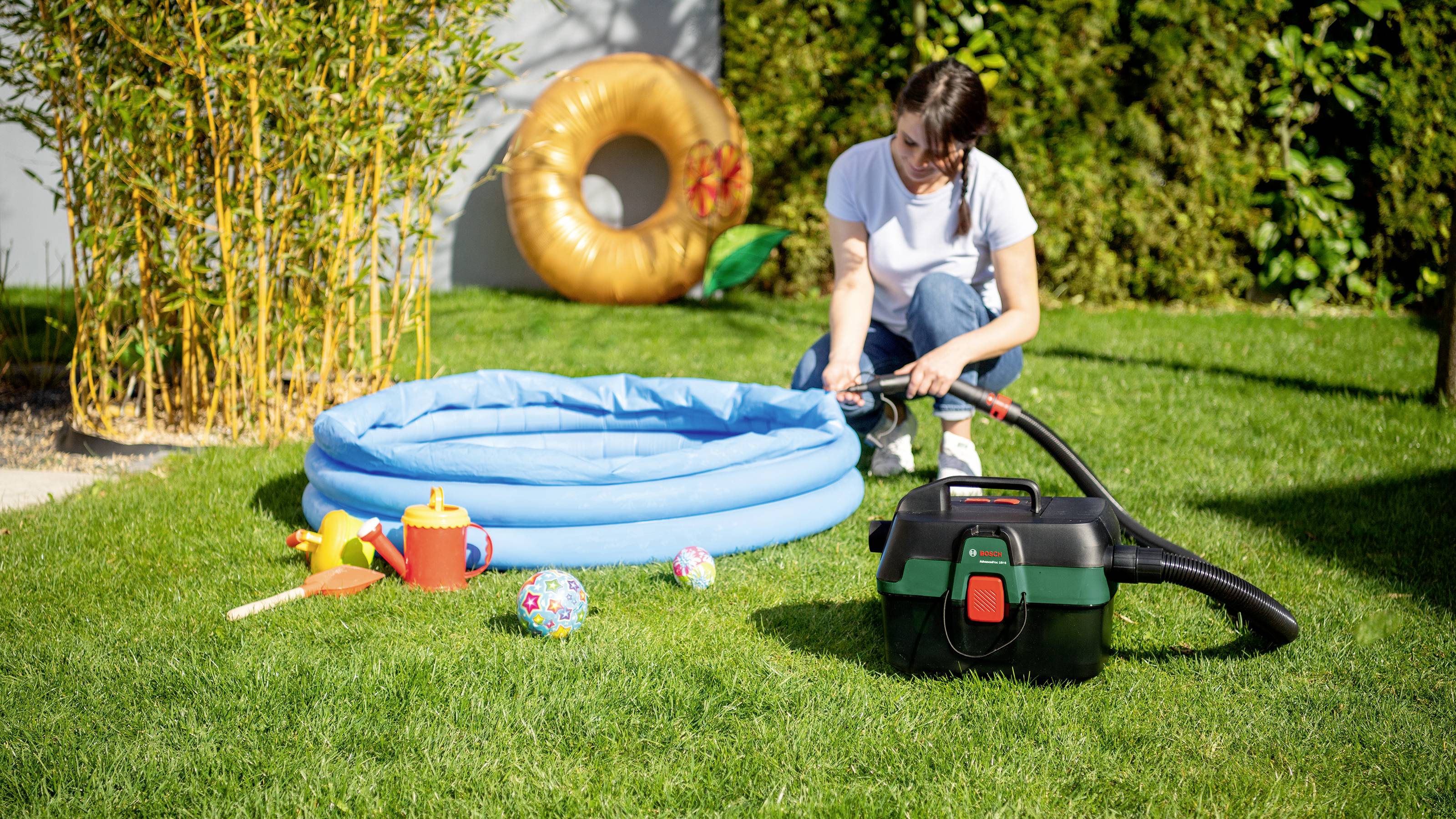 Une femme remplit une petite piscine bleue dans le jardin. Des jouets et un aspirateur sont placés à côté sur la pelouse.