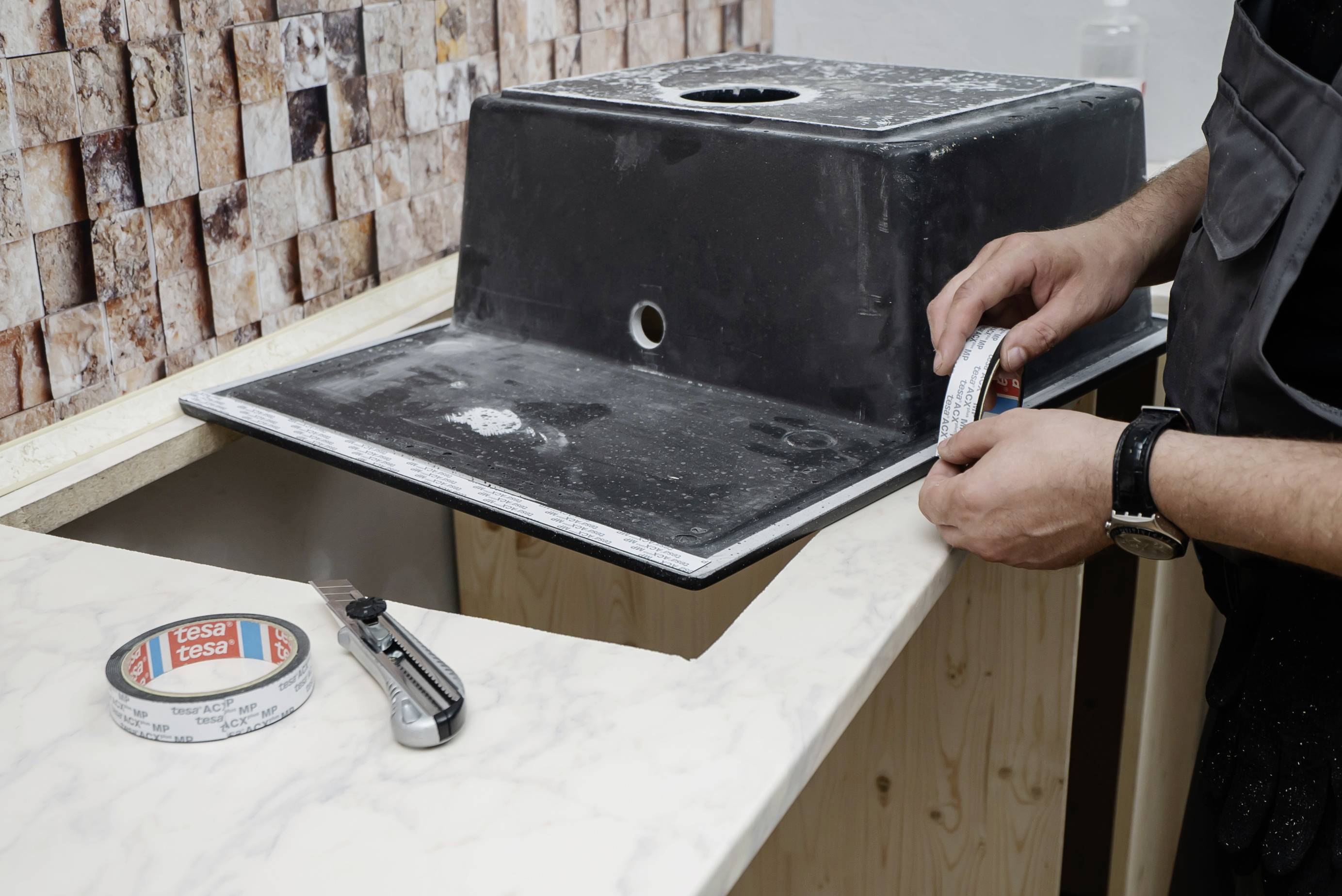 Une personne installe un lavabo dans un plan de travail. Des outils et du ruban d'étanchéité sont visibles.