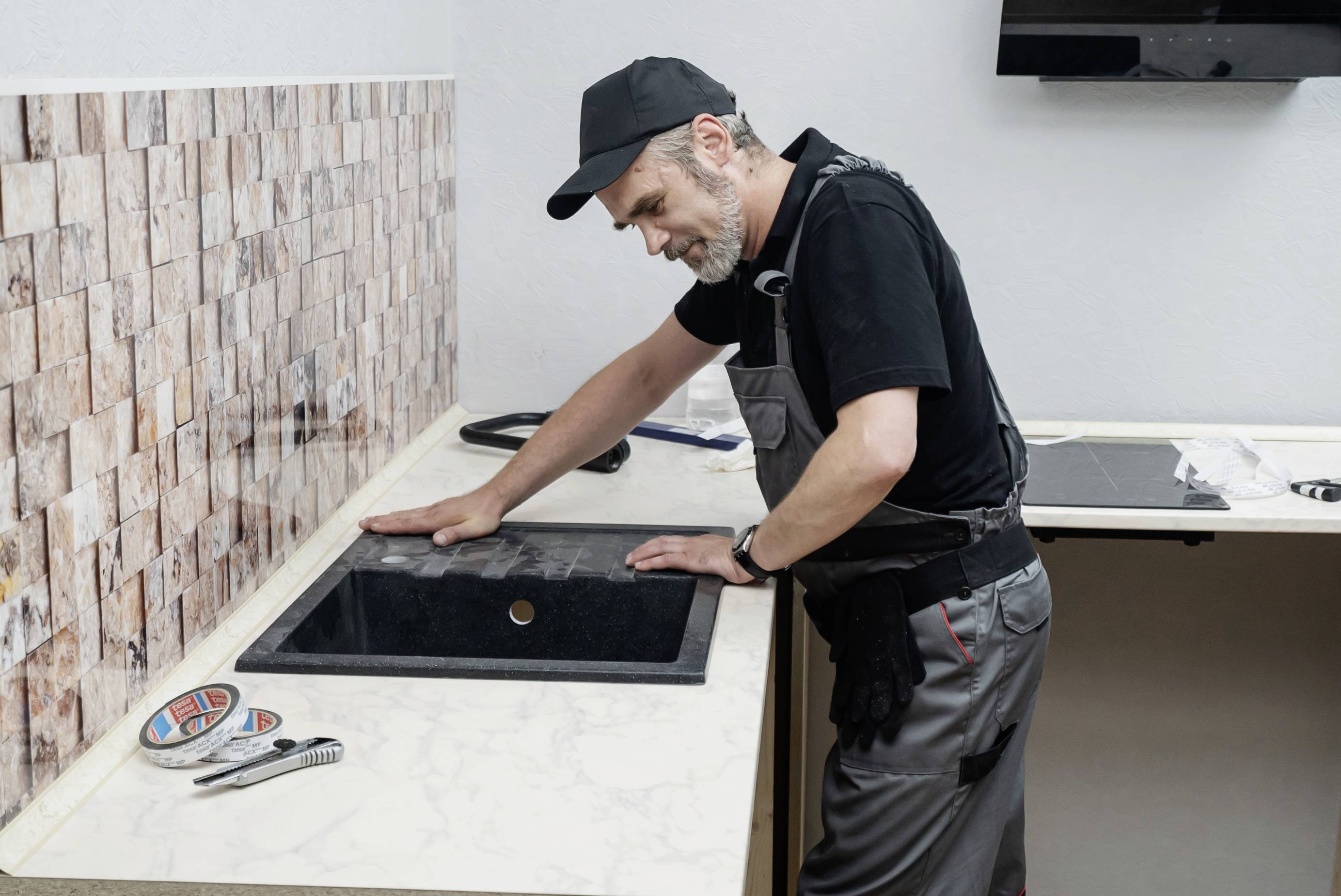 Un homme en tenue de travail vérifie l'installation d'un lavabo noir sur un plan de travail de cuisine.