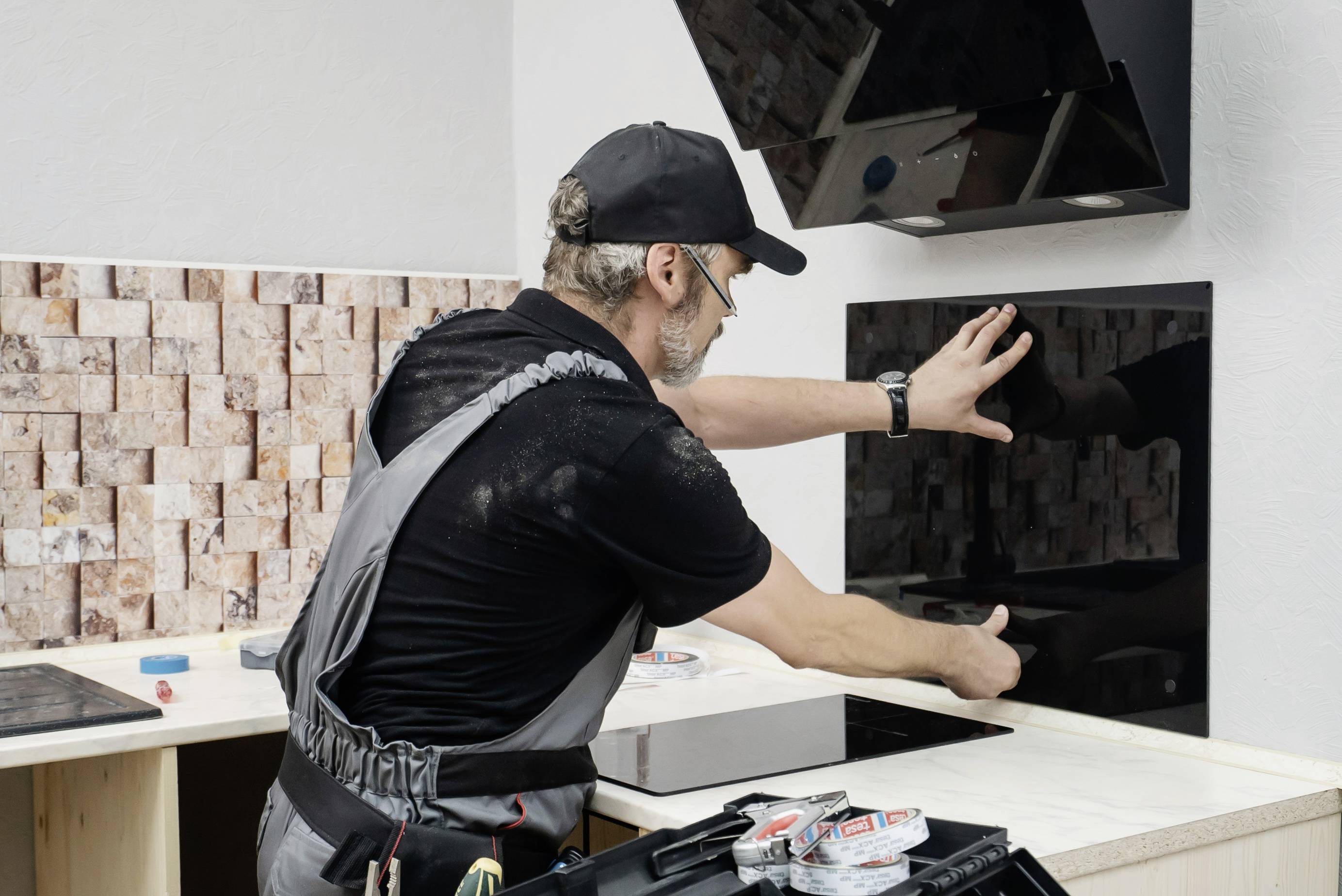 Un artisan installe une plaque de verre sur un mur de cuisine. Il porte des vêtements de travail et travaille avec précaution.