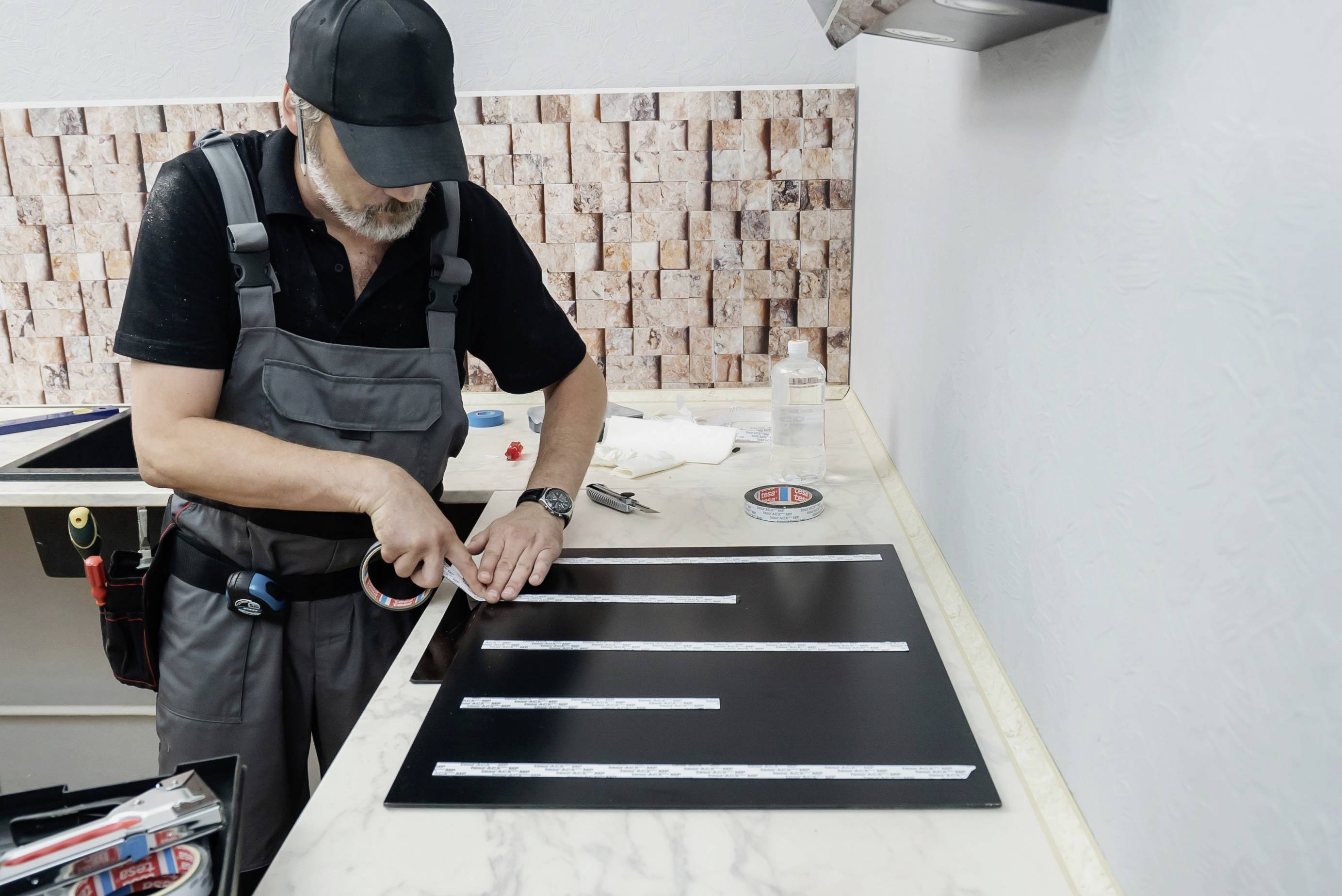 Un artisan en tenue de travail grise et casquette noire colle des bandes réfléchissantes sur un panneau noir posé sur une table.