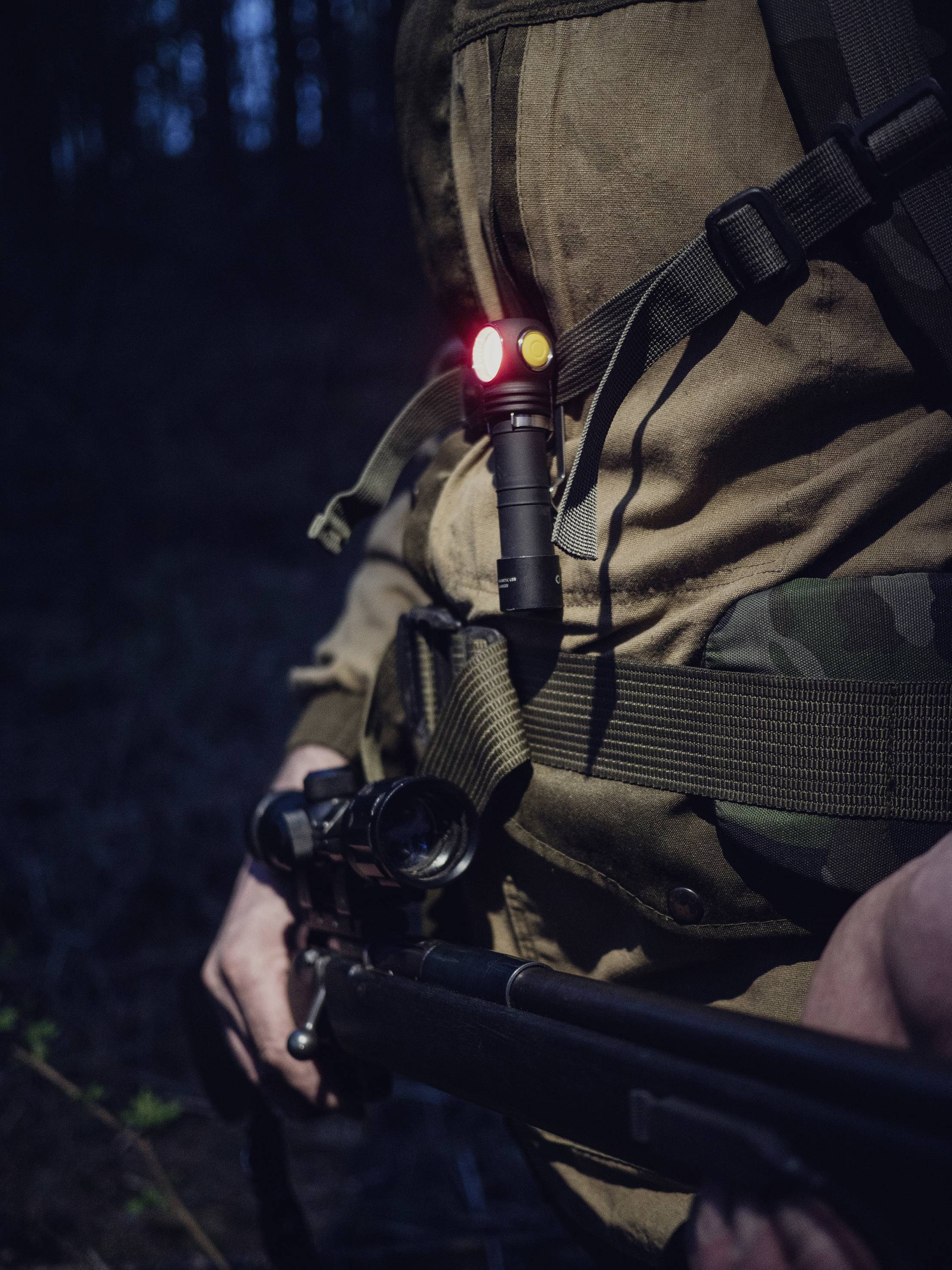 Une personne portant un uniforme de camouflage tient un fusil au crépuscule, avec une lampe de poche fixée sur la poitrine, émettant une lumière rouge et jaune.