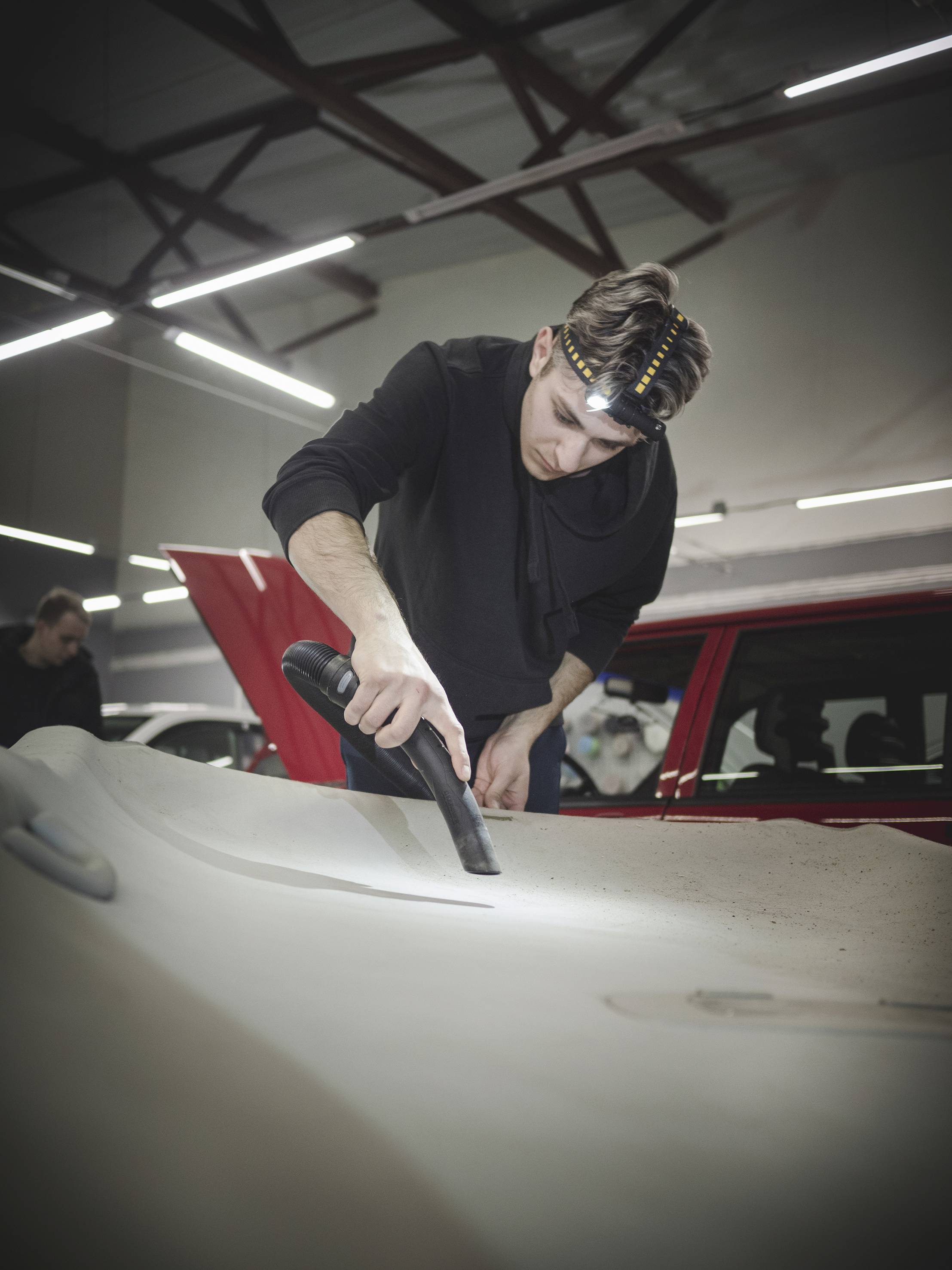 Un homme portant un bandeau sur la tête passe l'aspirateur sur le capot d'une voiture dans un atelier. À l'arrière-plan, on peut voir d'autres personnes et une voiture rouge.