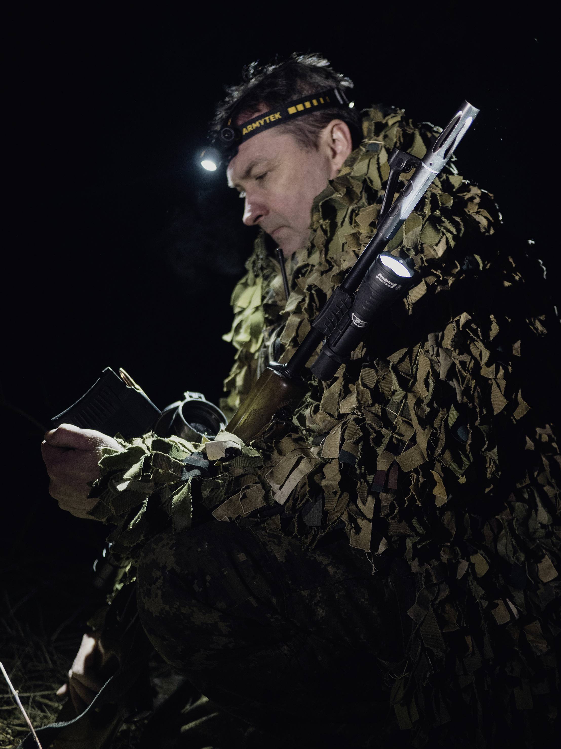 Une personne en tenue de camouflage et avec un bandeau frontal est assise dans l'obscurité, tenant un fusil, semblant concentrée et prête à agir.