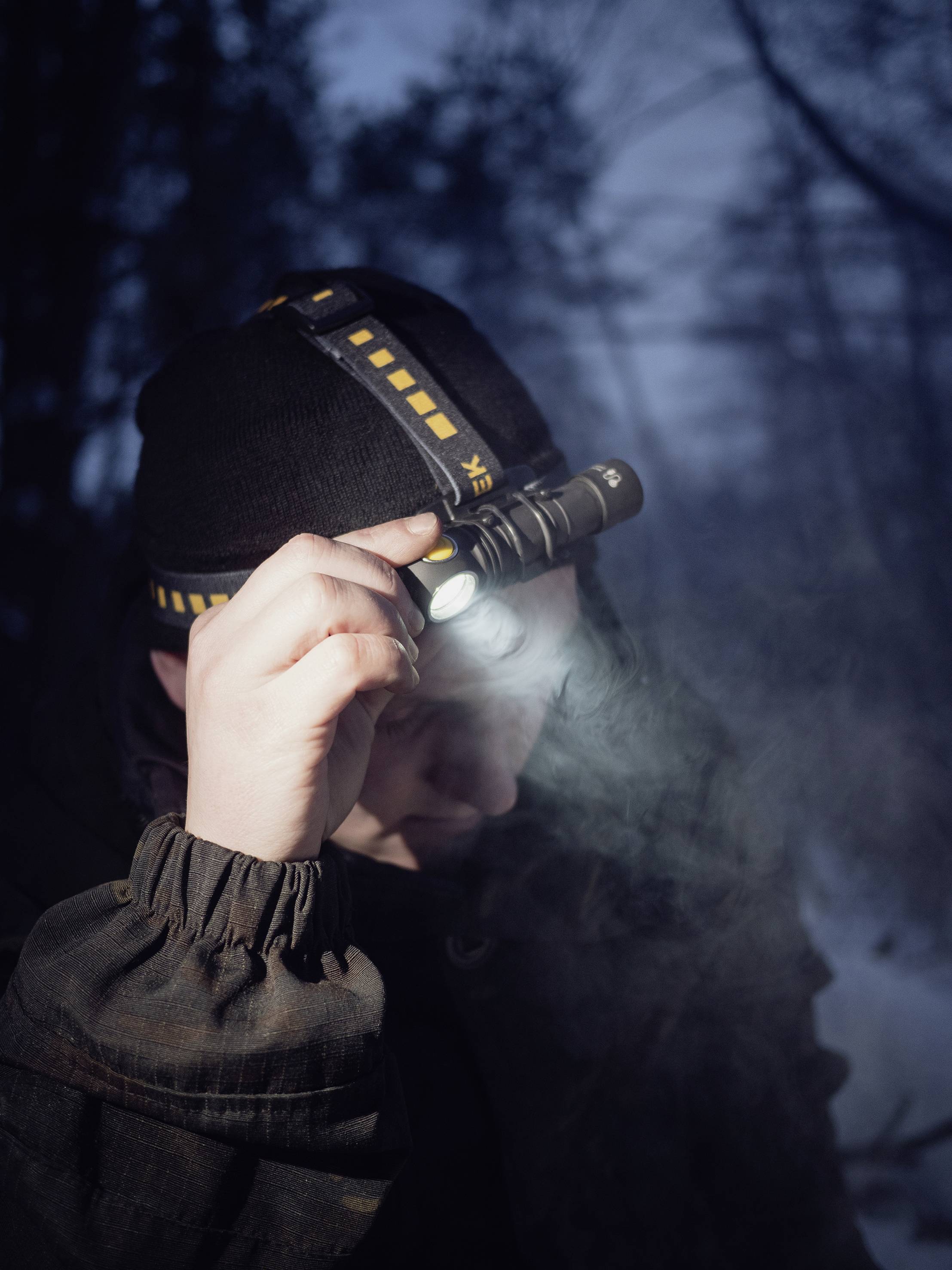 Une personne dans la forêt, vêtue de vêtements sombres, tient une lampe frontale lumineuse sur son front, regardant dans le crépuscule hivernal.