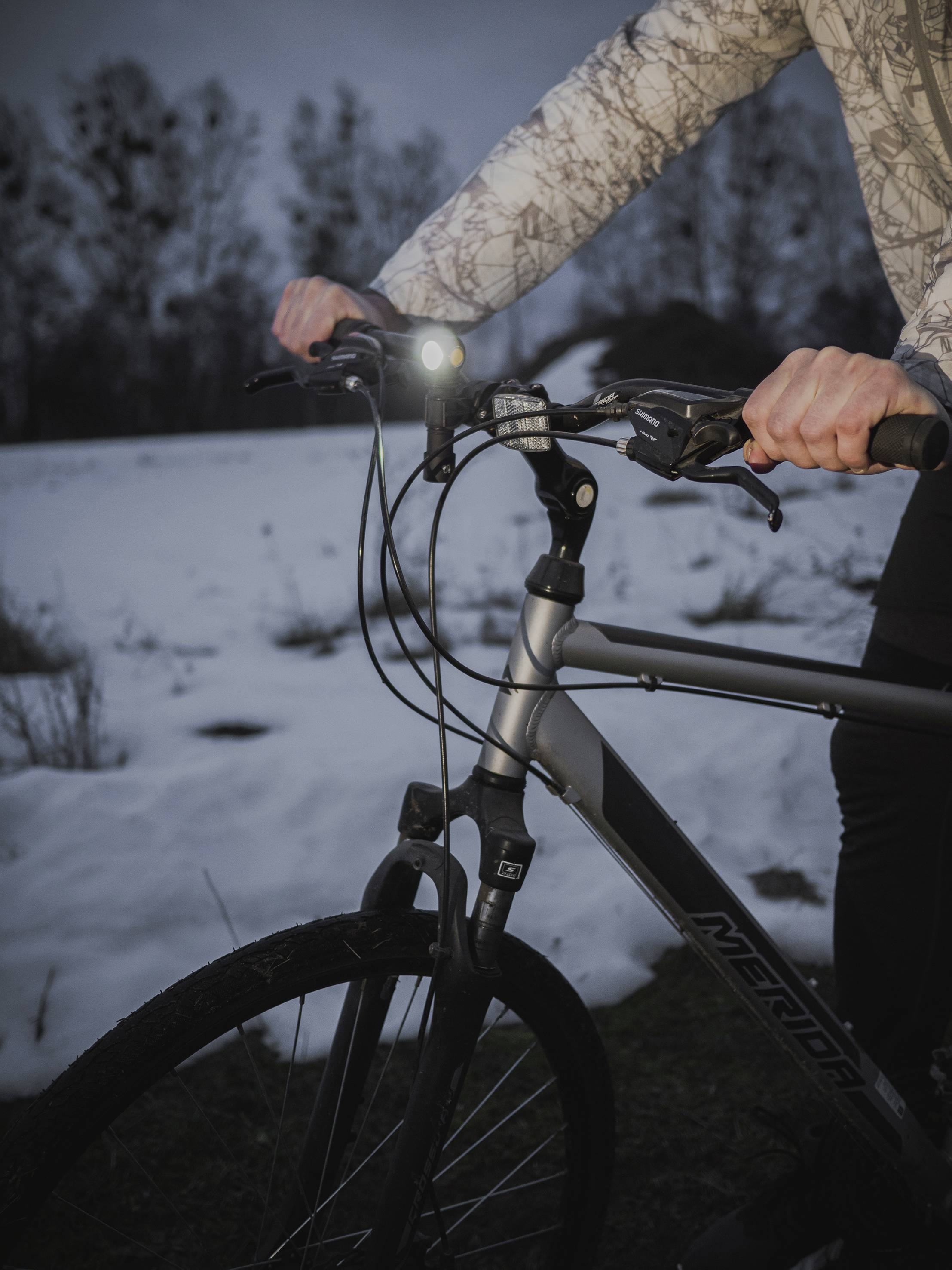 Une personne tient le guidon d'un vélo tout-terrain à l'extérieur. De la neige recouvre le sol, et le crépuscule s'installe. La lumière du vélo brille.