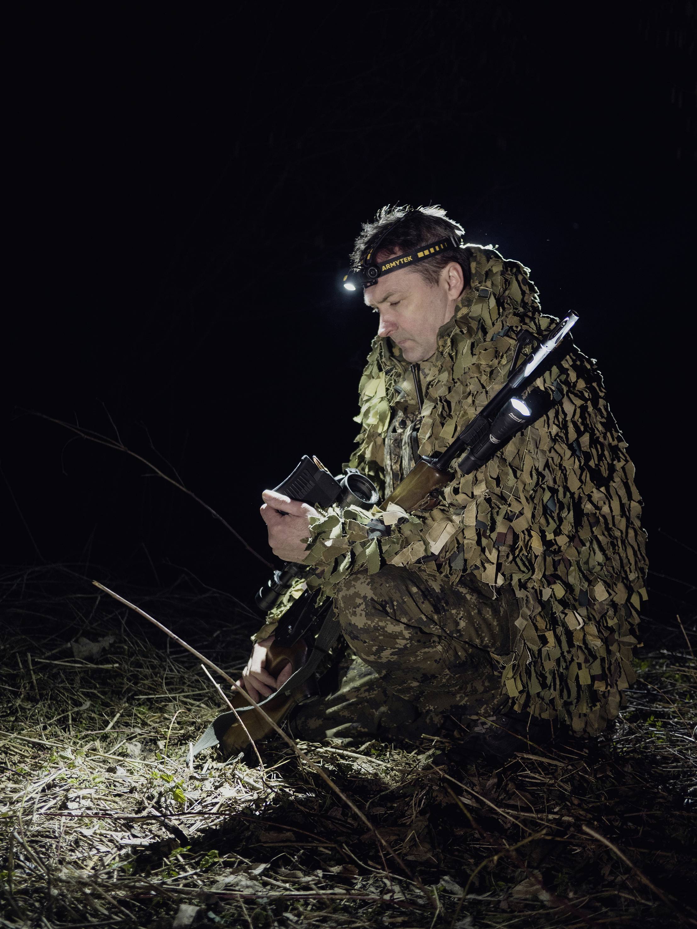 Une personne en tenue de camouflage est accroupie dehors la nuit, équipée d'un fusil et d'une lampe frontale, lisant ou vérifiant quelque chose dans sa main.