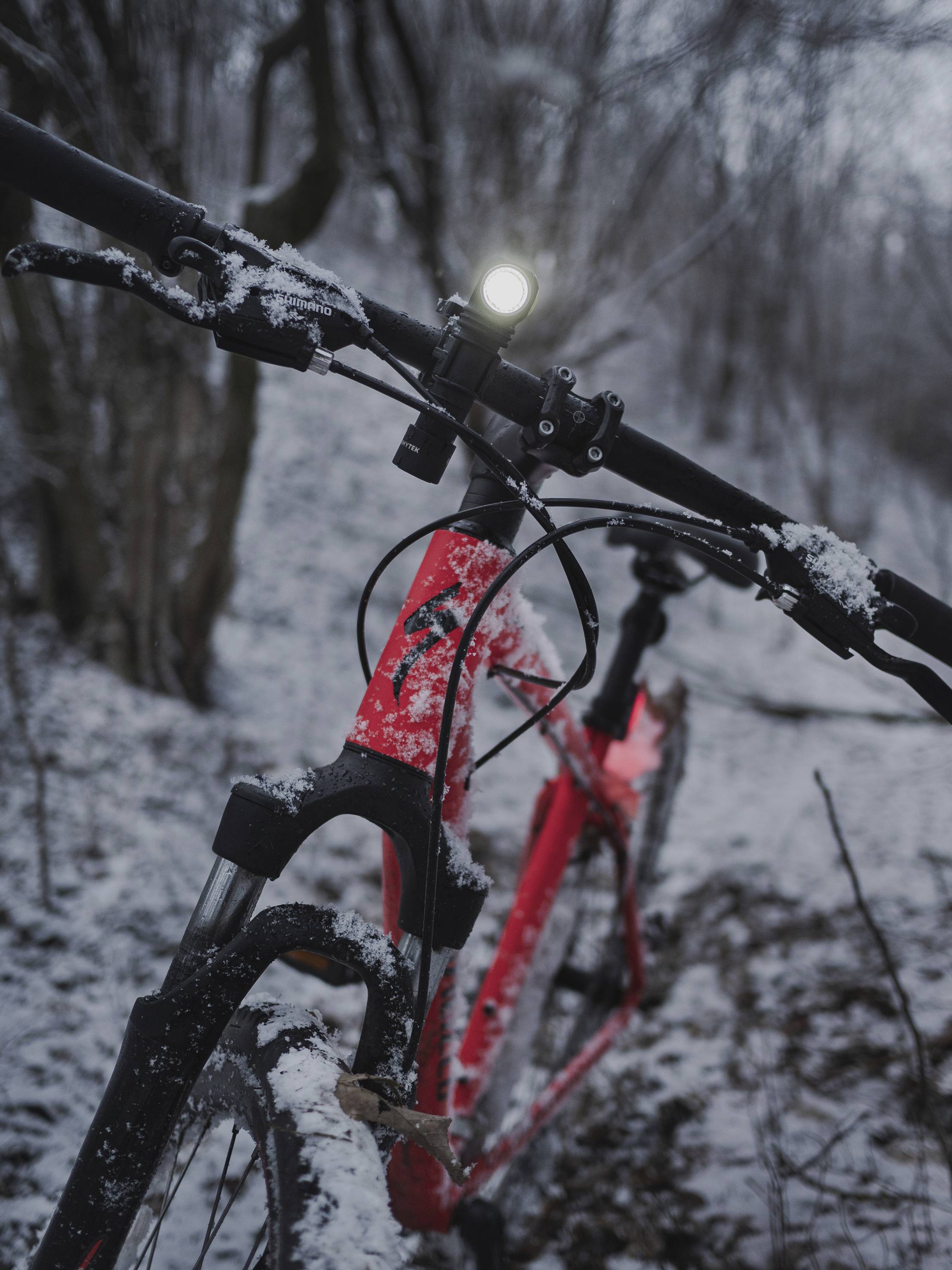 Vélo rouge avec un phare avant allumé sur un chemin forestier enneigé au crépuscule. Les pneus et le cadre sont recouverts de neige.