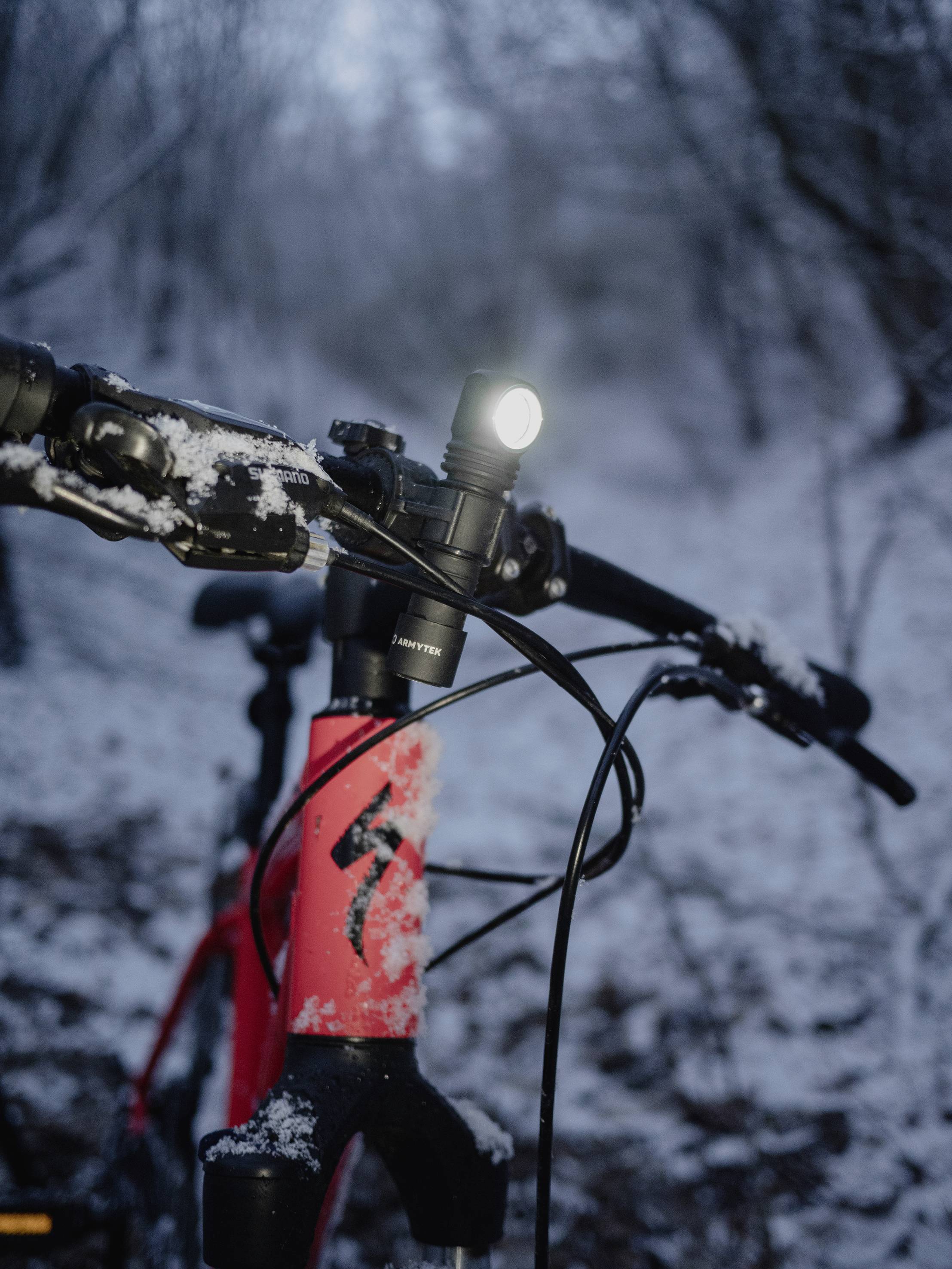 Gros plan d'un vélo rouge avec un phare avant allumé, sur un chemin enneigé en forêt au crépuscule. Le guidon est partiellement recouvert de neige.