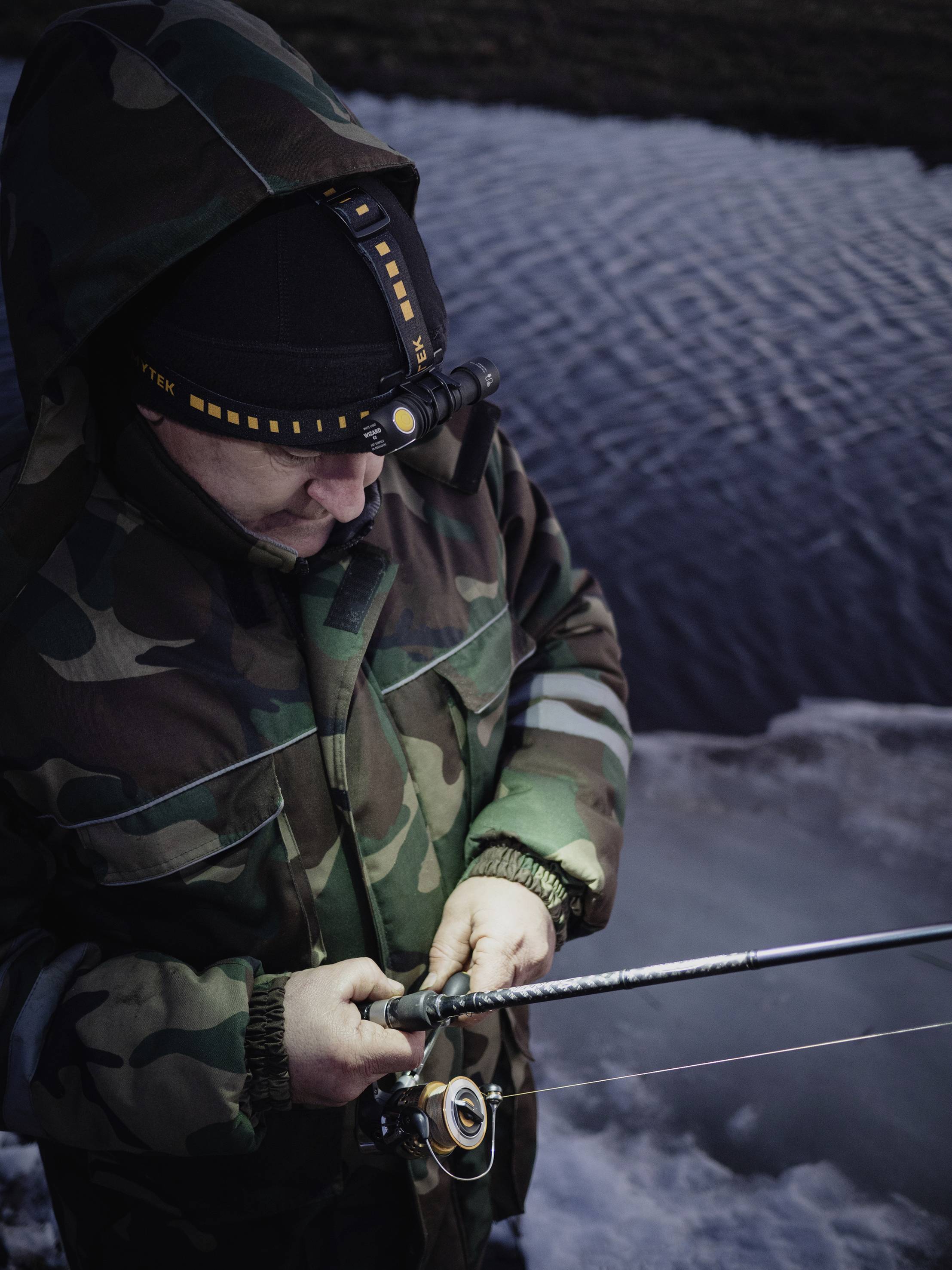 Une personne en tenue de camouflage pêche près de l'eau. C'est l'hiver, et la zone au bord de l'eau est gelée. La personne est concentrée sur sa canne à pêche.