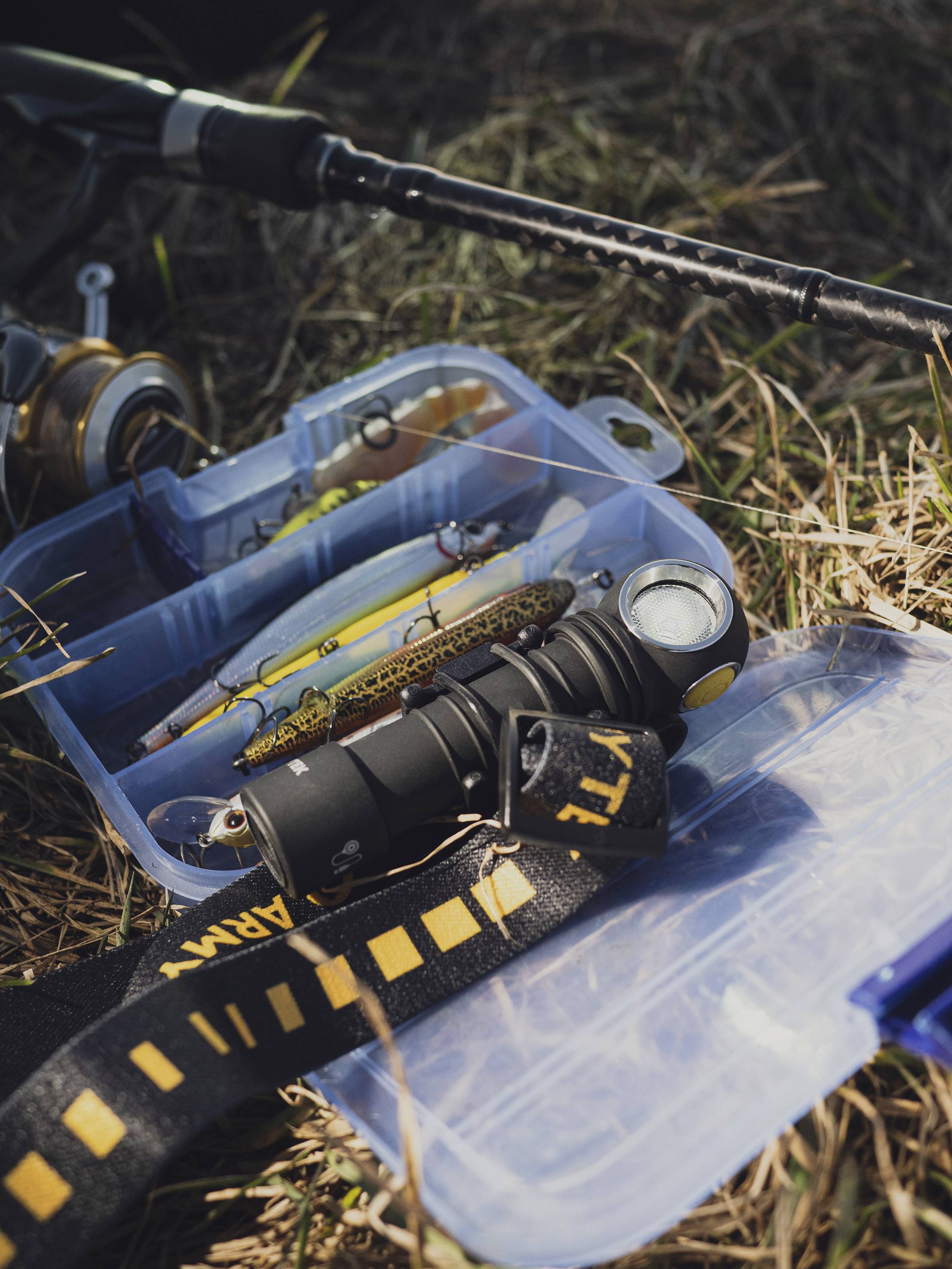 Accessoires de pêche sur un sol herbeux, avec une boîte à pêche, des appâts et une canne à pêche, prêts pour une sortie de pêche.