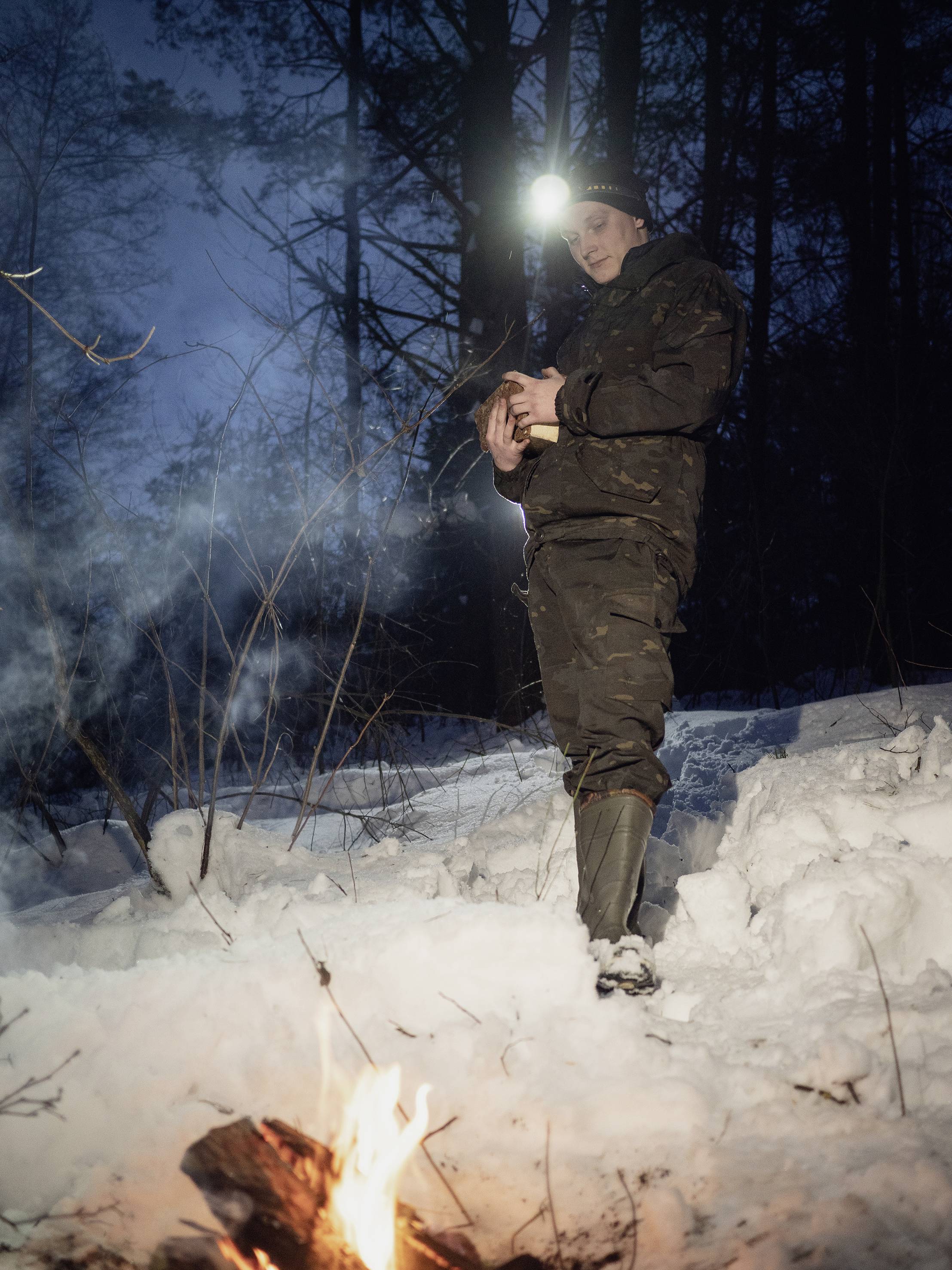 Une personne vêtue de vêtements d'hiver se tient dans la neige en forêt, tenant un animal dans ses bras. Au premier plan, un petit feu de camp brûle.