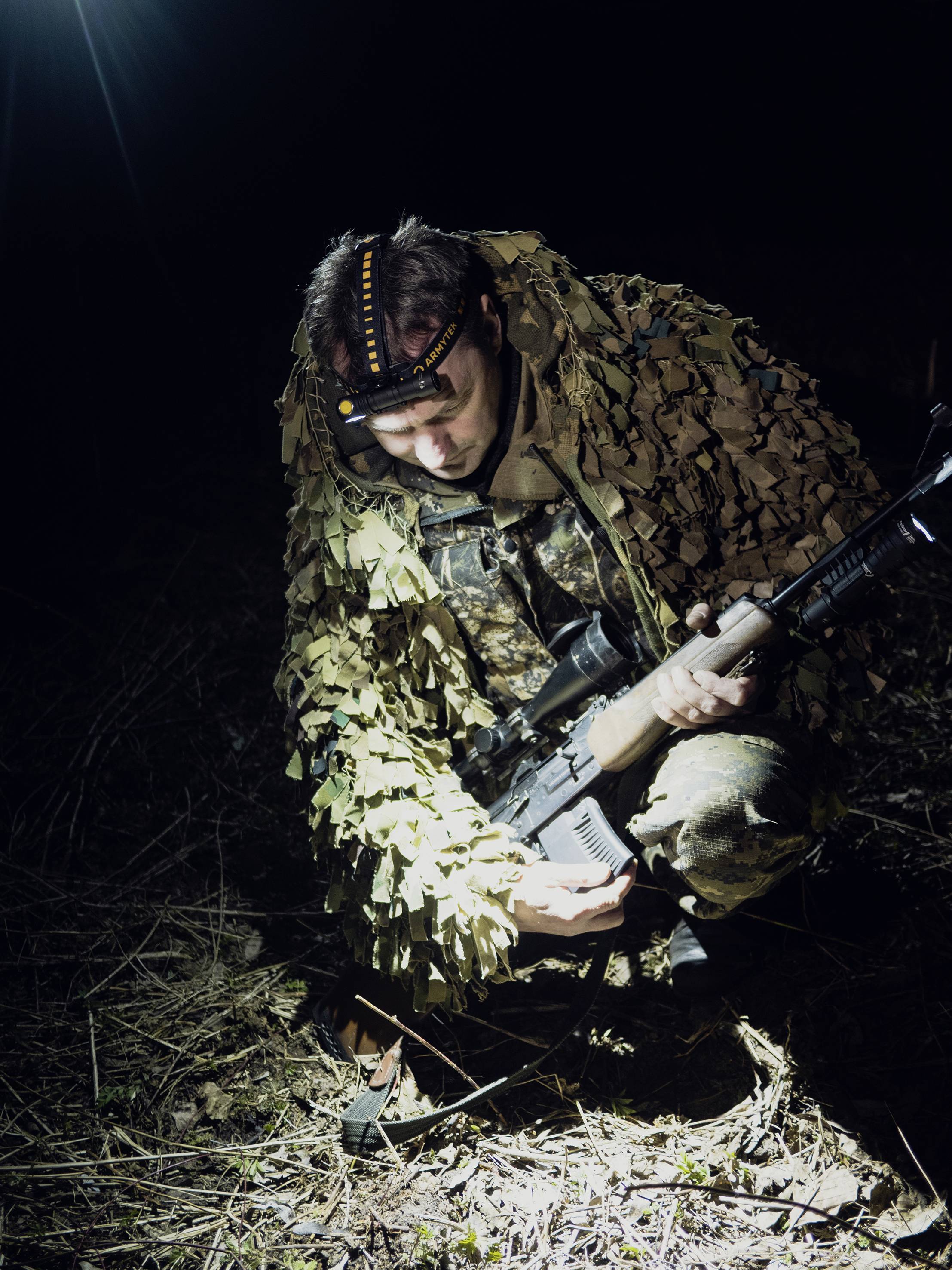 Un homme en tenue de camouflage est accroupi au sol la nuit, tenant un fusil dans les deux mains et éclairé d'en haut.