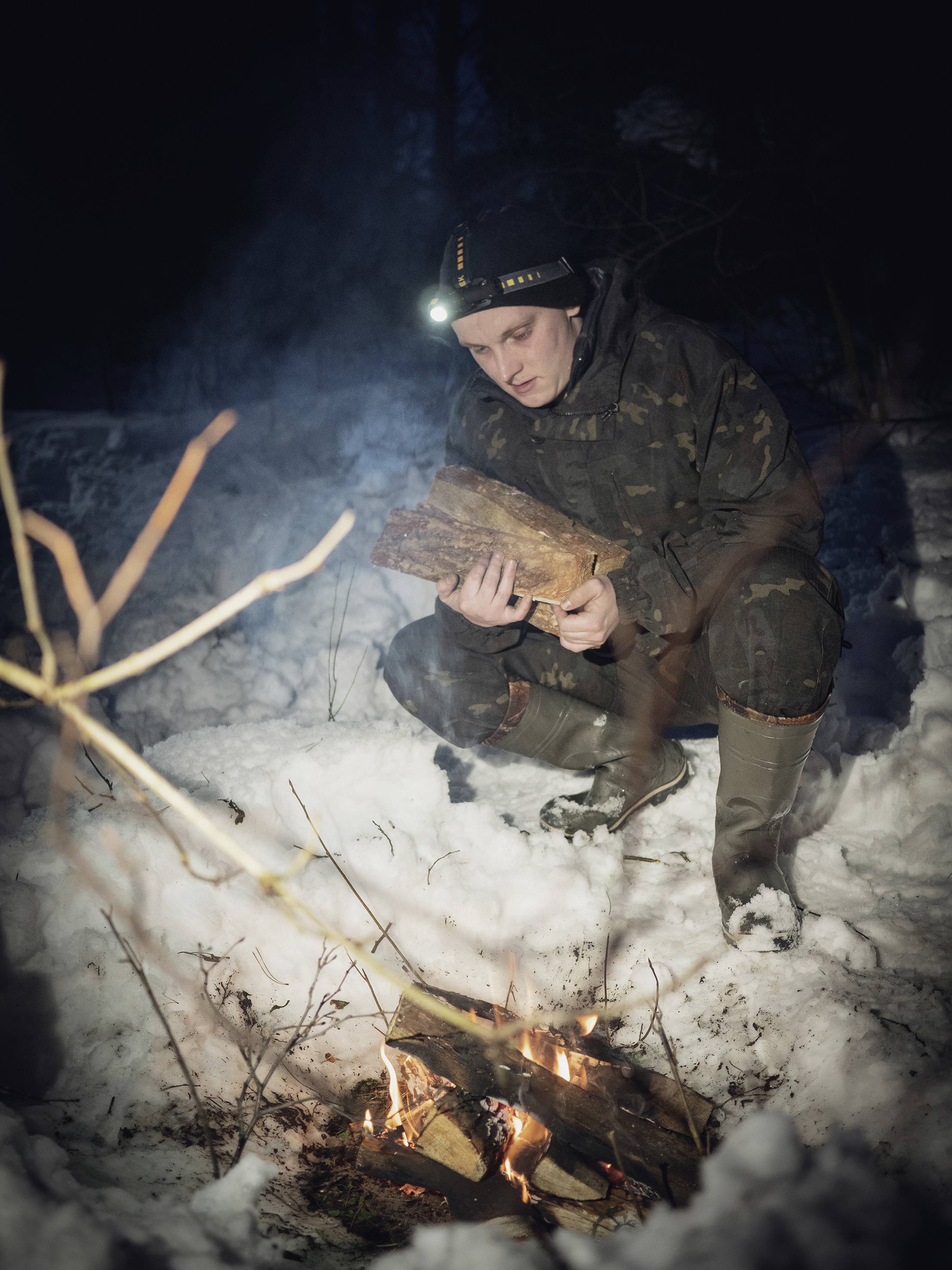 Une personne dans la forêt dispose des bûches sur un feu de camp dans la neige. Elle porte un bandeau frontal et des vêtements chauds.