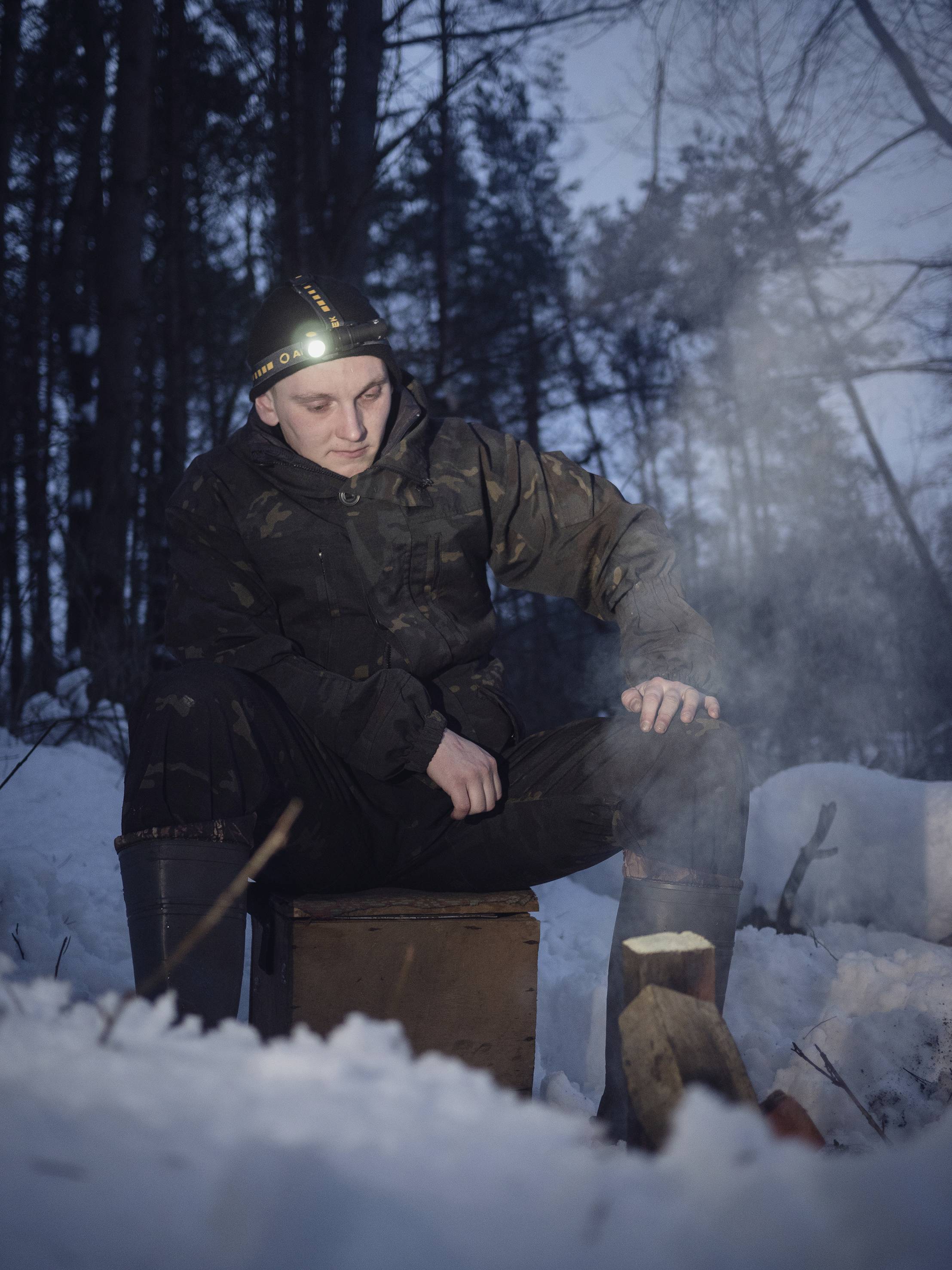 Une personne est assise dans des vêtements d'hiver sombres et portant une lampe frontale sur un caisson en bois dans la forêt, entourée de neige. De la fumée s'élève d'un petit feu.